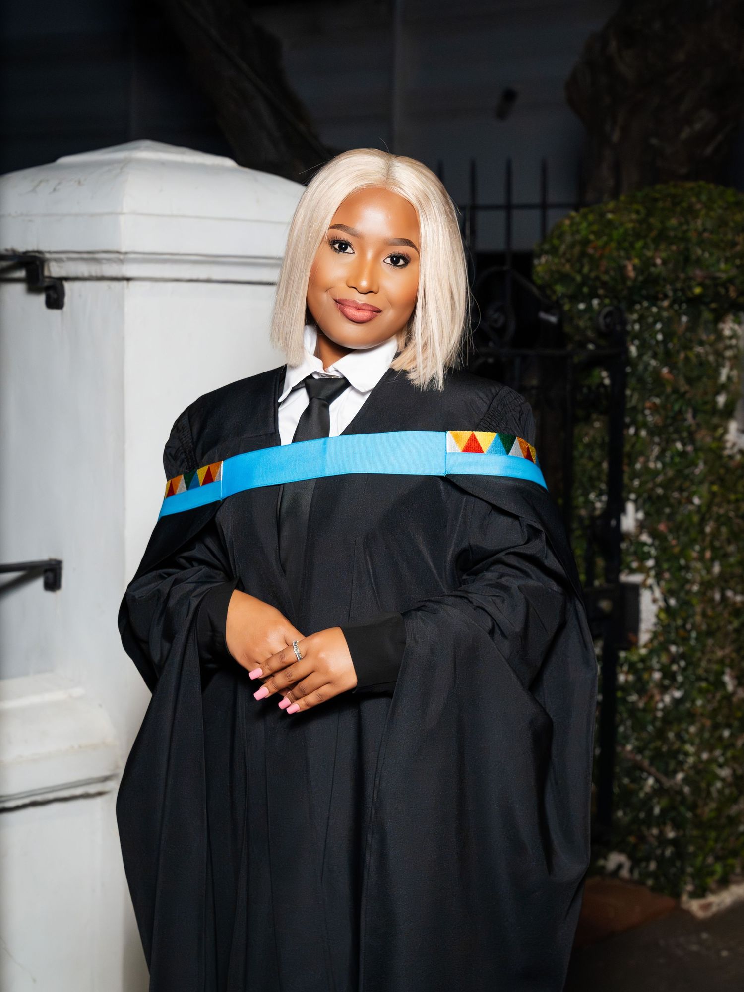 Young woman in black graduation gown with colorful sash and blonde bob hairstyle posing proudly outdoors.