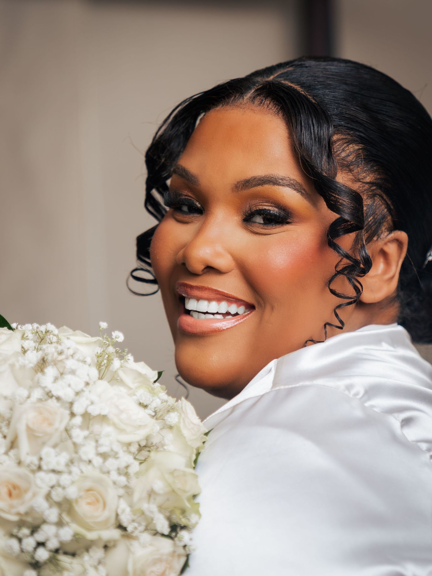 Smiling bride in white satin holding a bouquet of white roses and baby's breath, with elegant curled hair.
