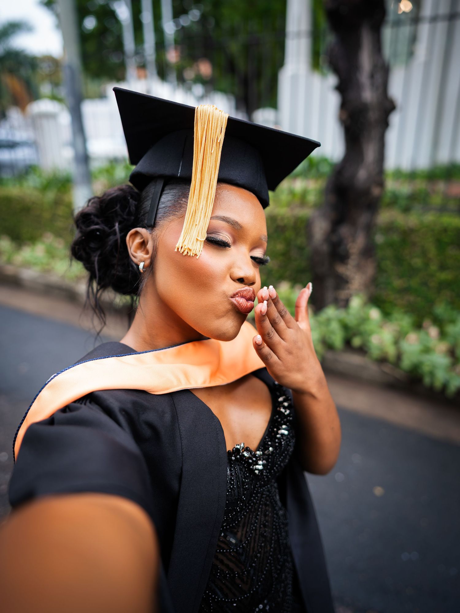 A young woman in black graduation gown and cap with gold tassel blowing a kiss during a selfie outdoors.