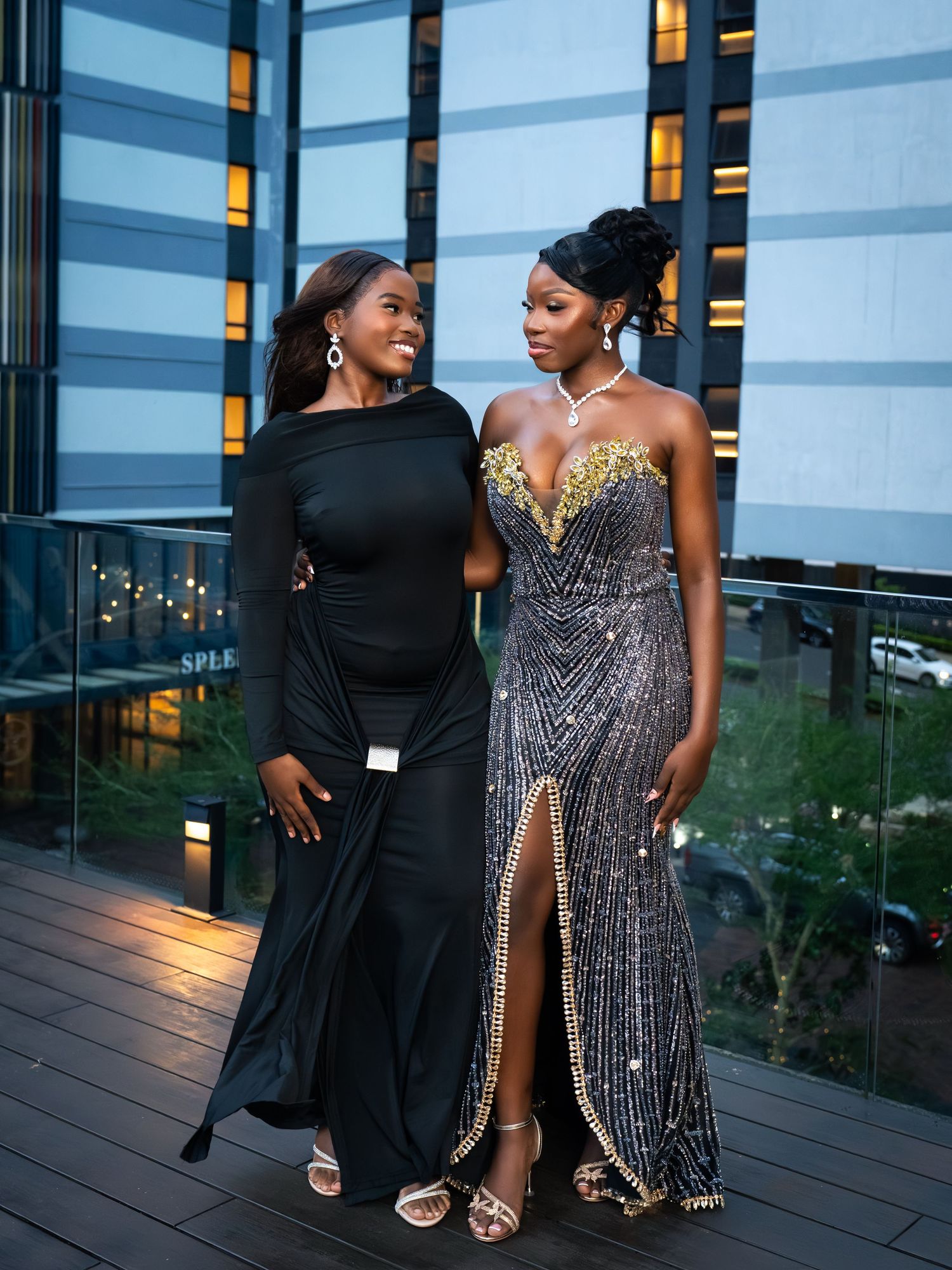Two elegant women in formal gowns pose on a rooftop terrace at dusk, one in black and one in sequined dress.