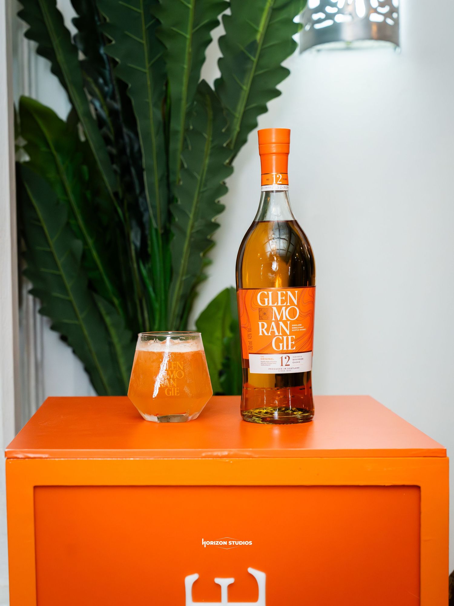 Glenmorangie 12 Year whisky bottle with a glass of amber cocktail on an orange cabinet with tropical plant backdrop.