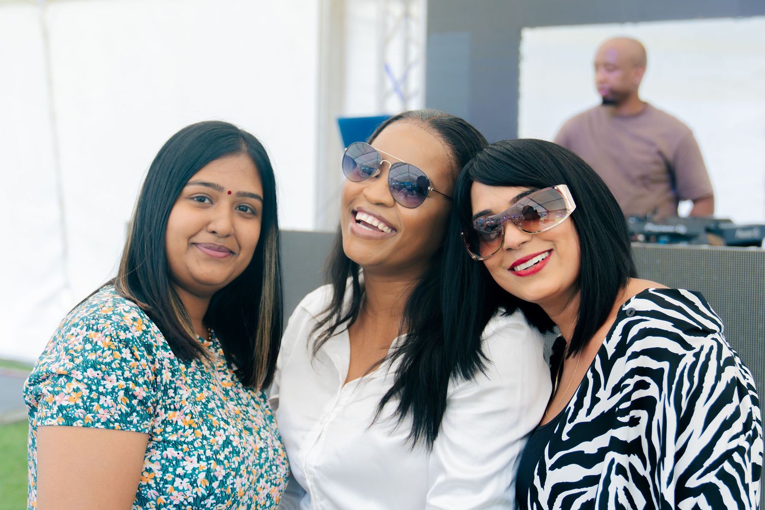 Three stylish women wearing sunglasses smiling together at an indoor event with a DJ in background.