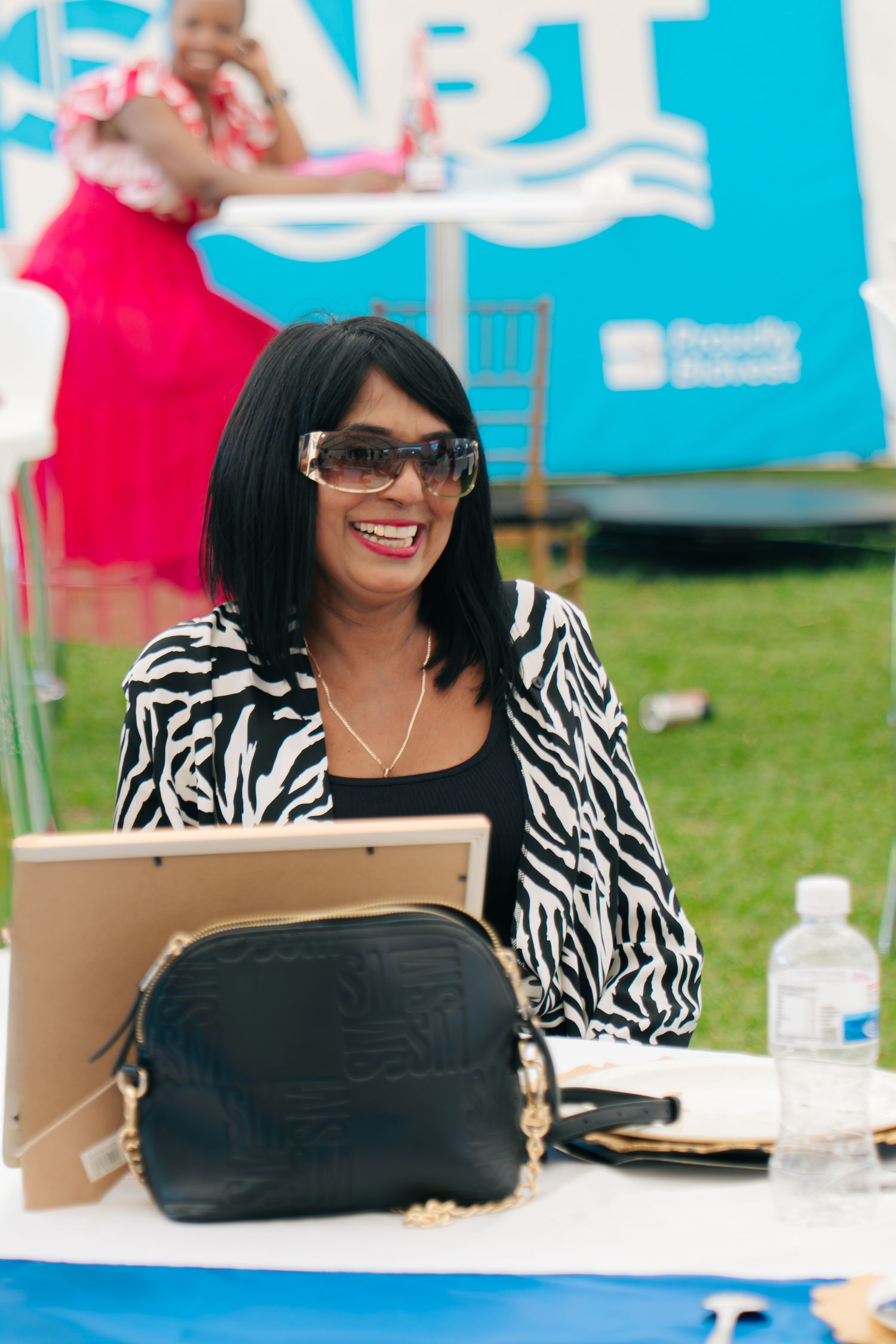 Smiling woman in zebra-print jacket and sunglasses sits at outdoor event table with black purse and tablet.