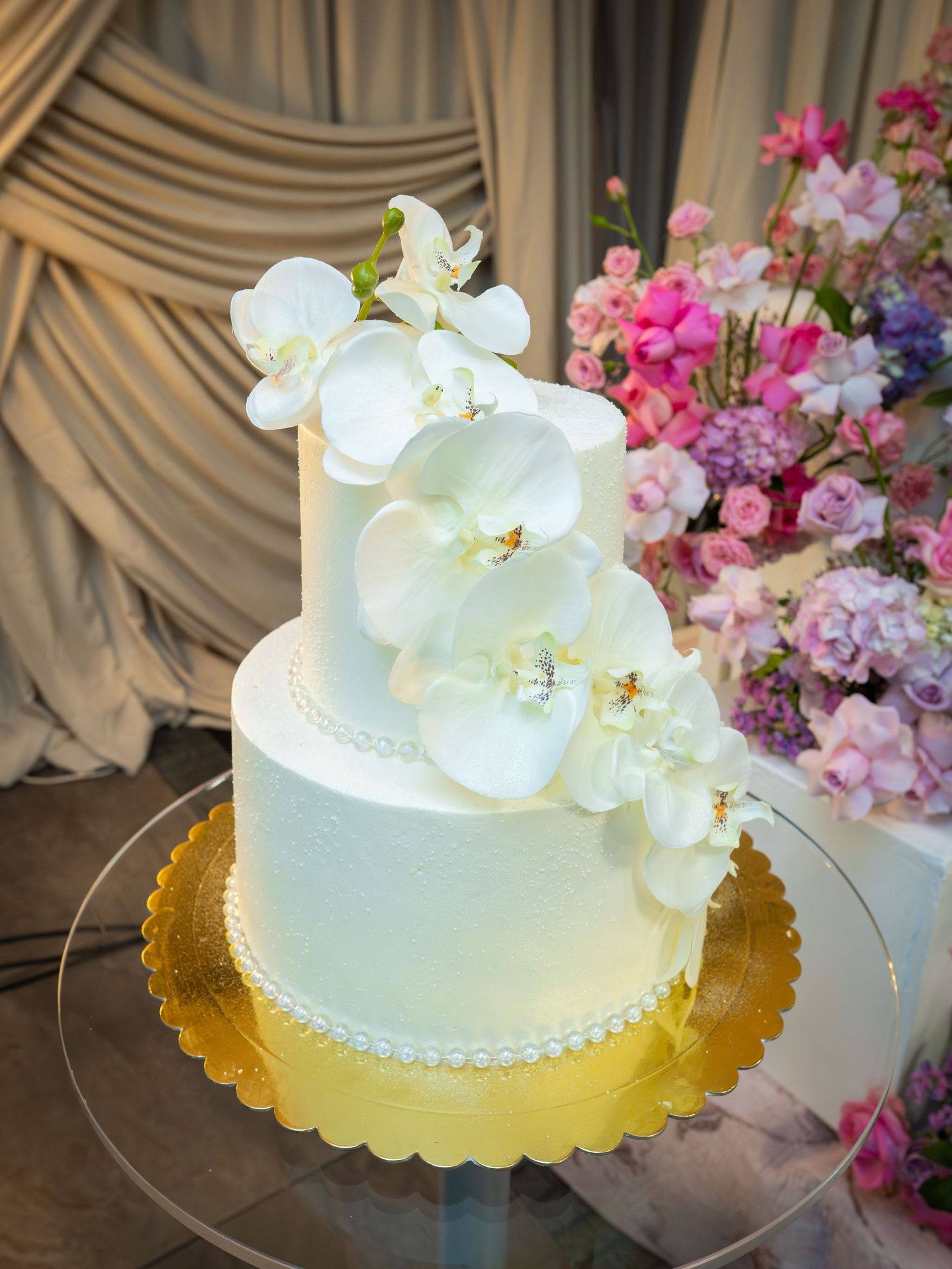 Elegant two-tier white wedding cake with white orchids on a gold board, surrounded by pink floral arrangements.