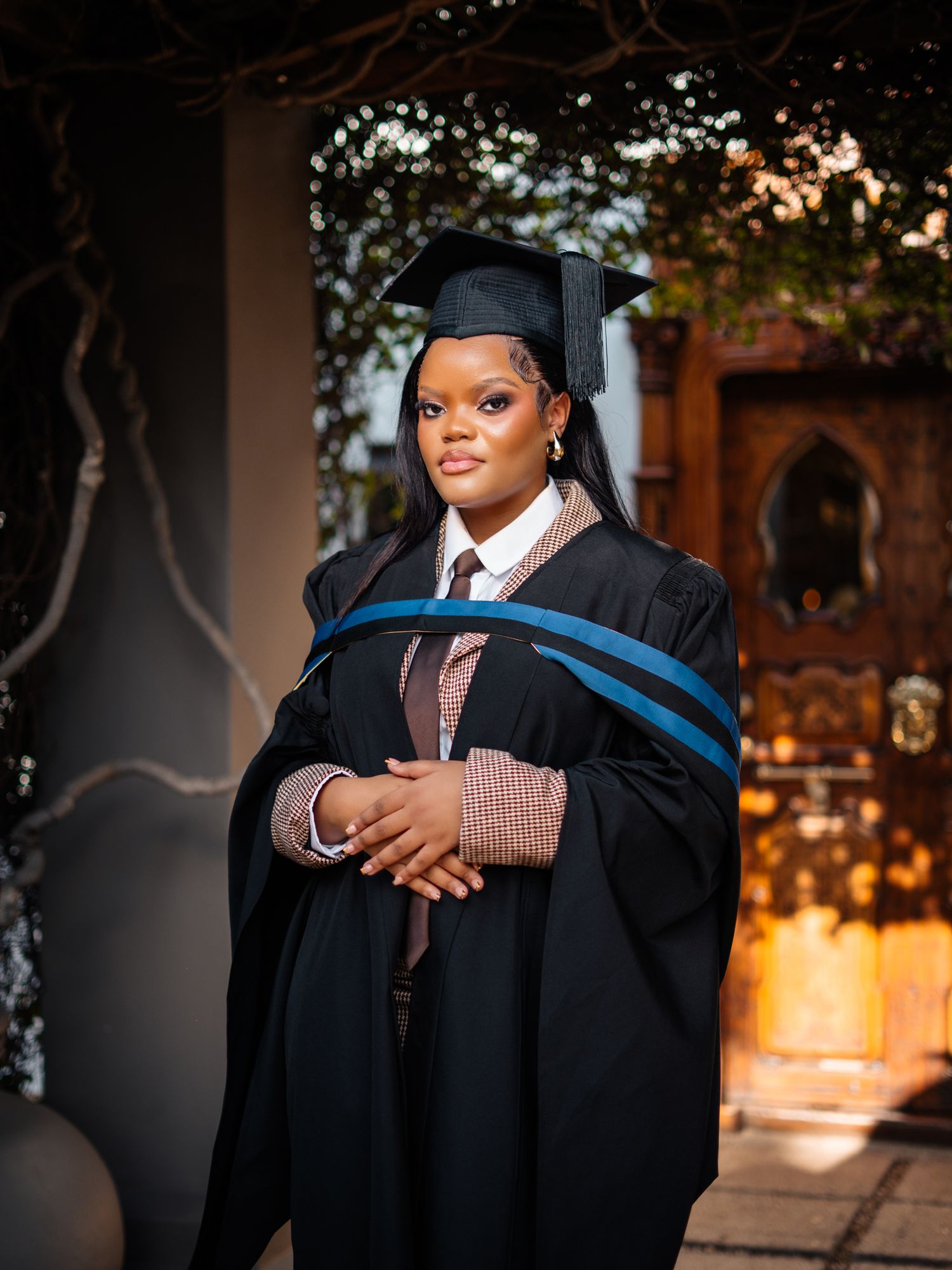 Young woman in black graduation gown and cap with blue sash poses proudly outdoors for graduation photo.