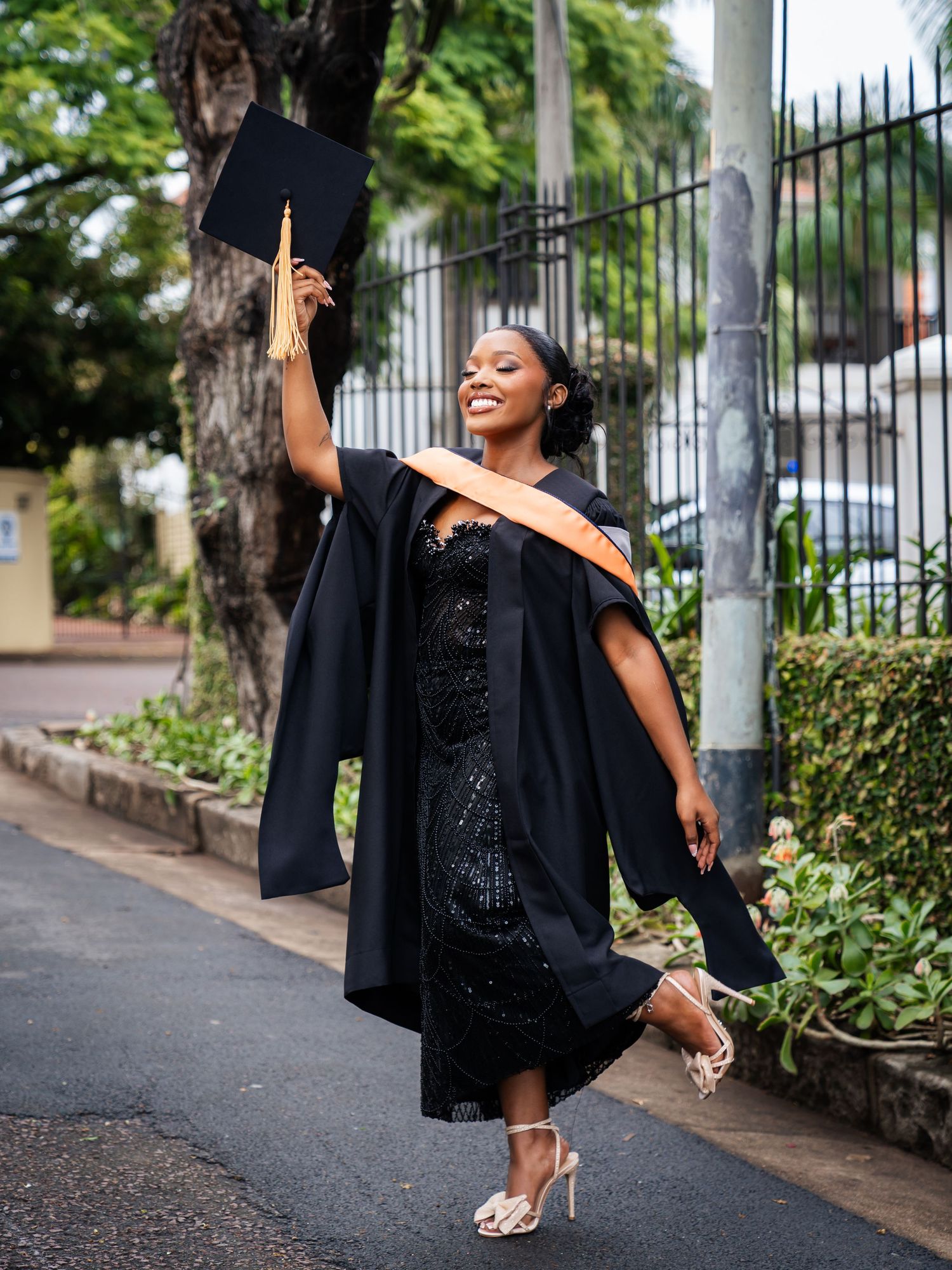Joyful graduate in black cap and gown celebrates outdoors, holding mortarboard aloft in a tree-lined street.