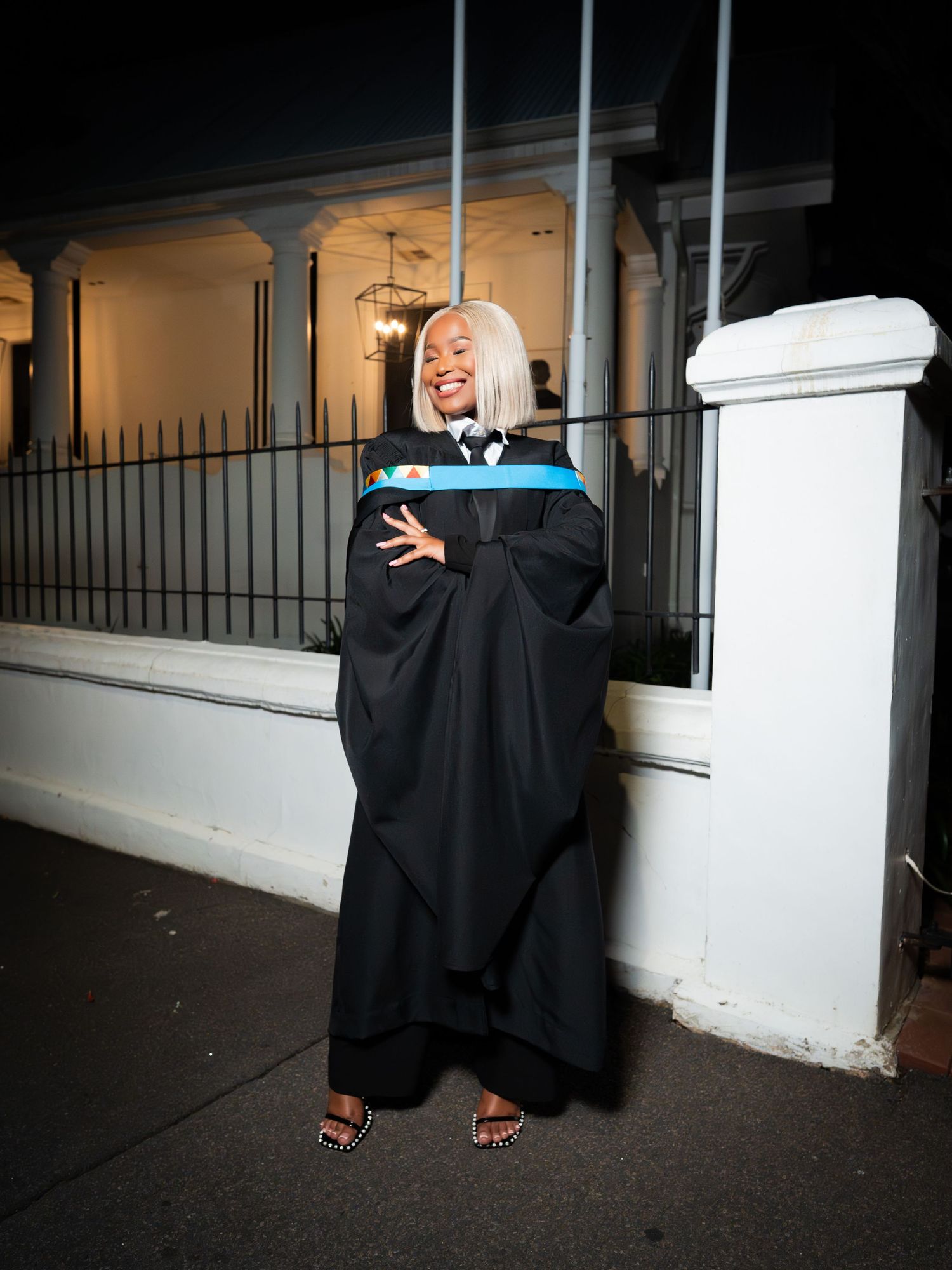 A graduate in black academic gown and blue hood poses proudly outside at night.