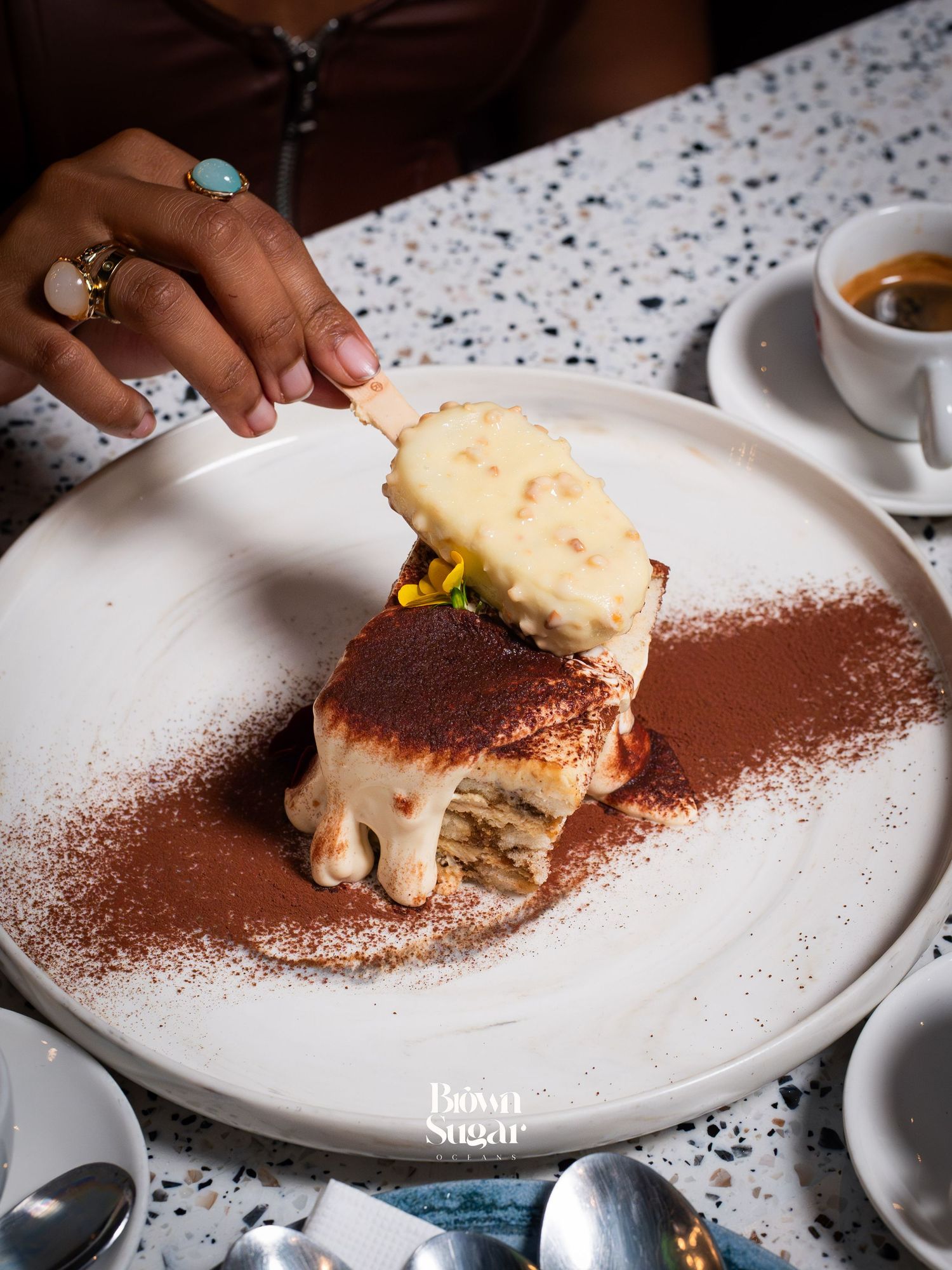 A hand lifts a slice of creamy tiramisu dusted with cocoa powder on a white plate at Brown Sugar.