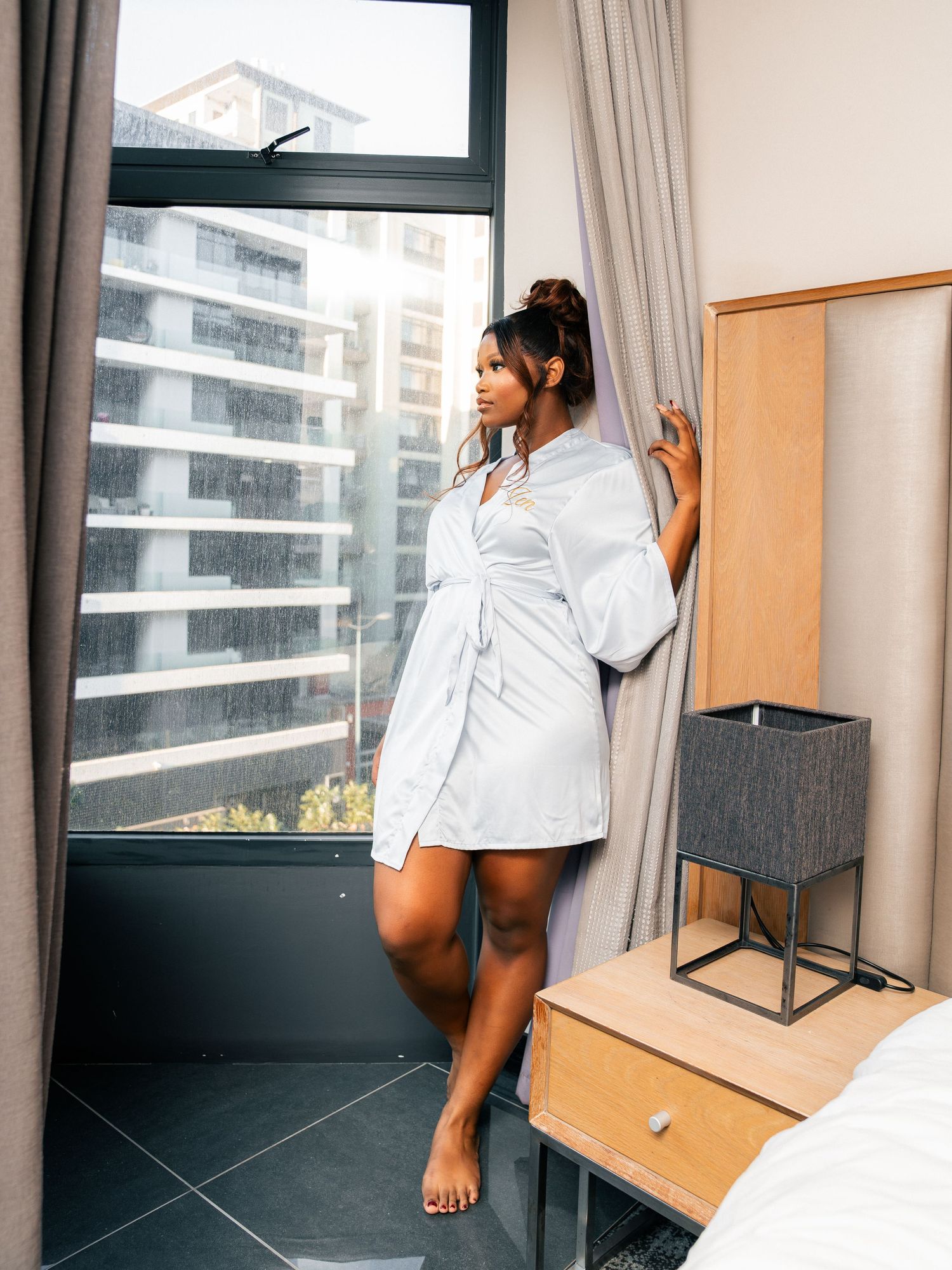 A woman in a white robe stands barefoot by a large window in a modern hotel room, gazing at the city view.
