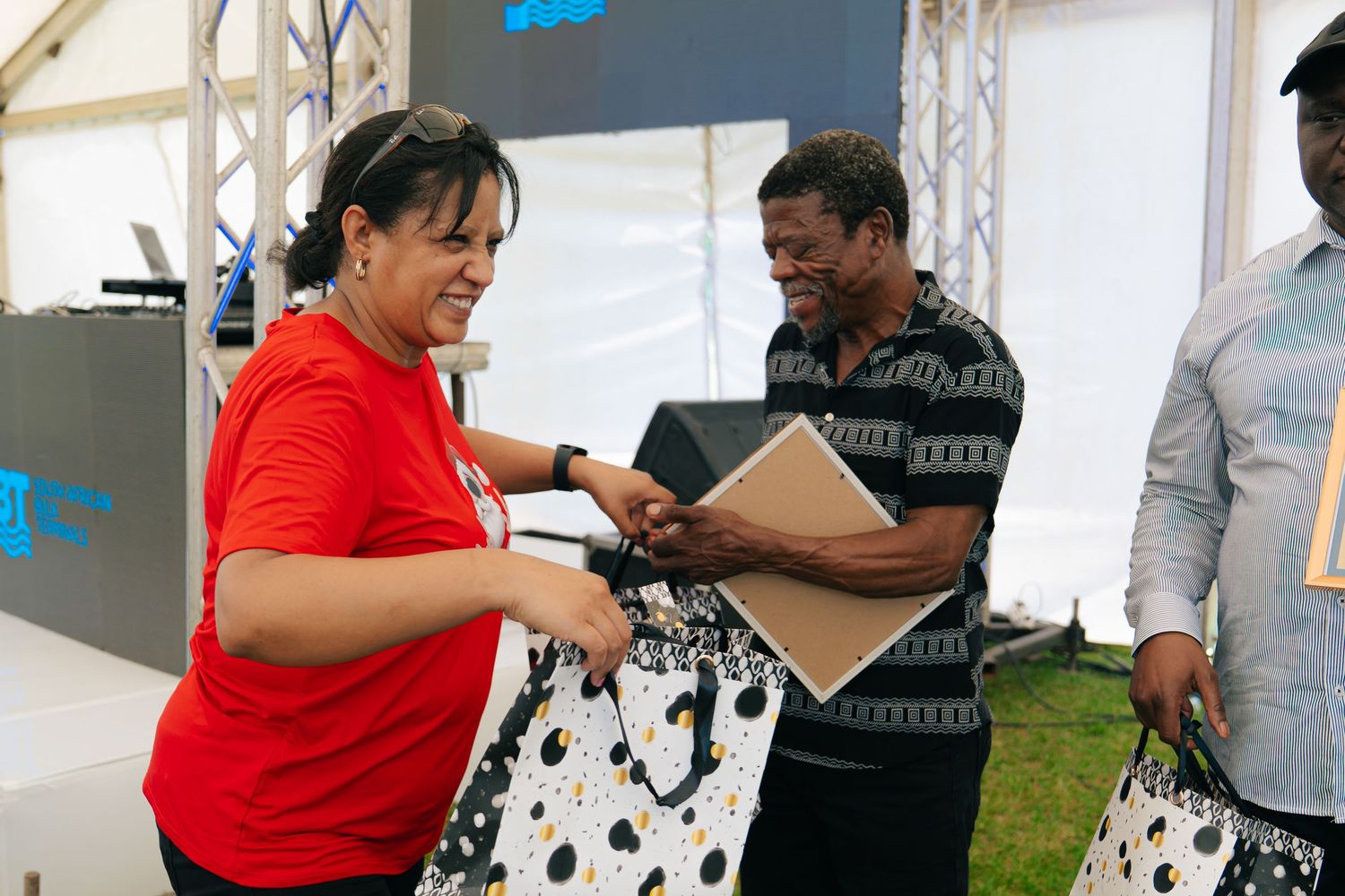 Two people exchanging gift bags at an outdoor corporate event under a white tent.
