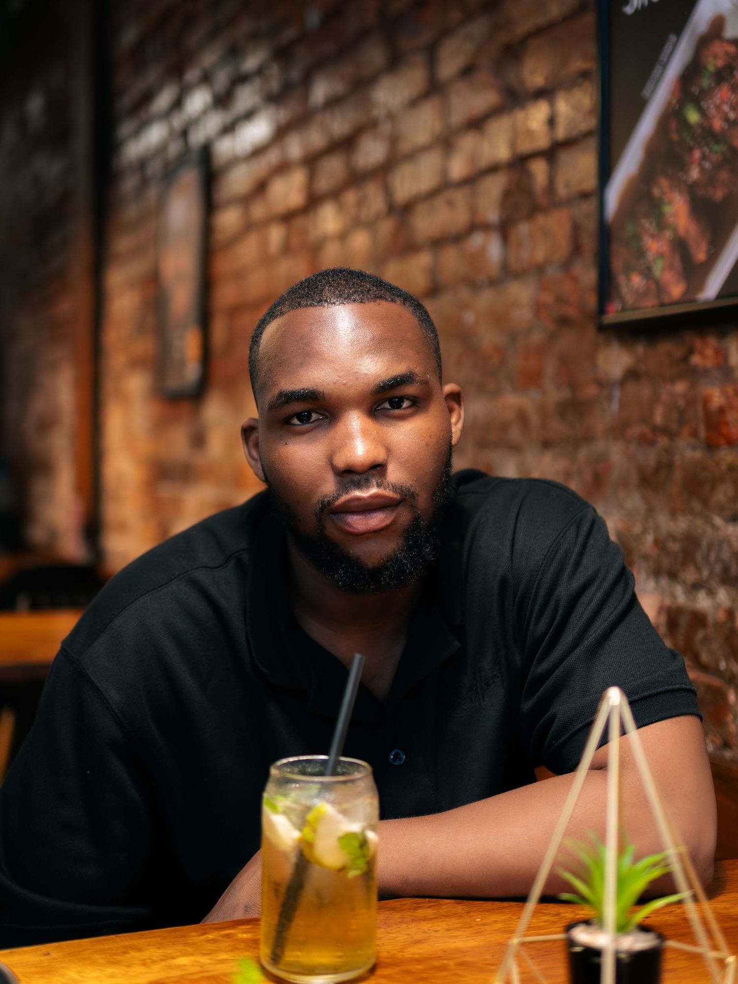 Young Black man in black shirt sits at wooden table with mojito cocktail in brick-walled restaurant.