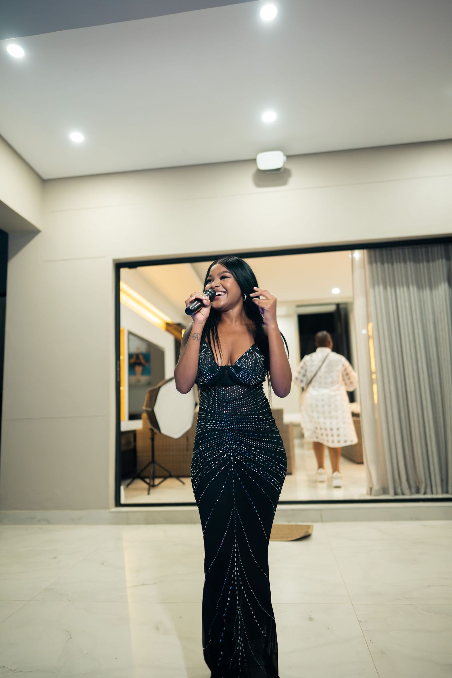 A woman in a black sequined gown singing into a microphone at an elegant indoor event.