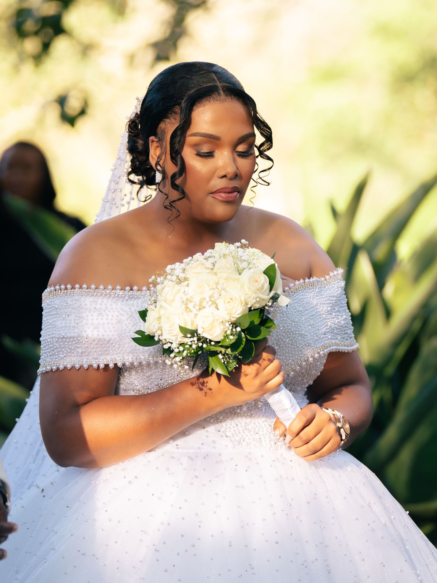 A bride in an off-shoulder pearl wedding gown holds a white rose bouquet, wearing a veil with curled hair.