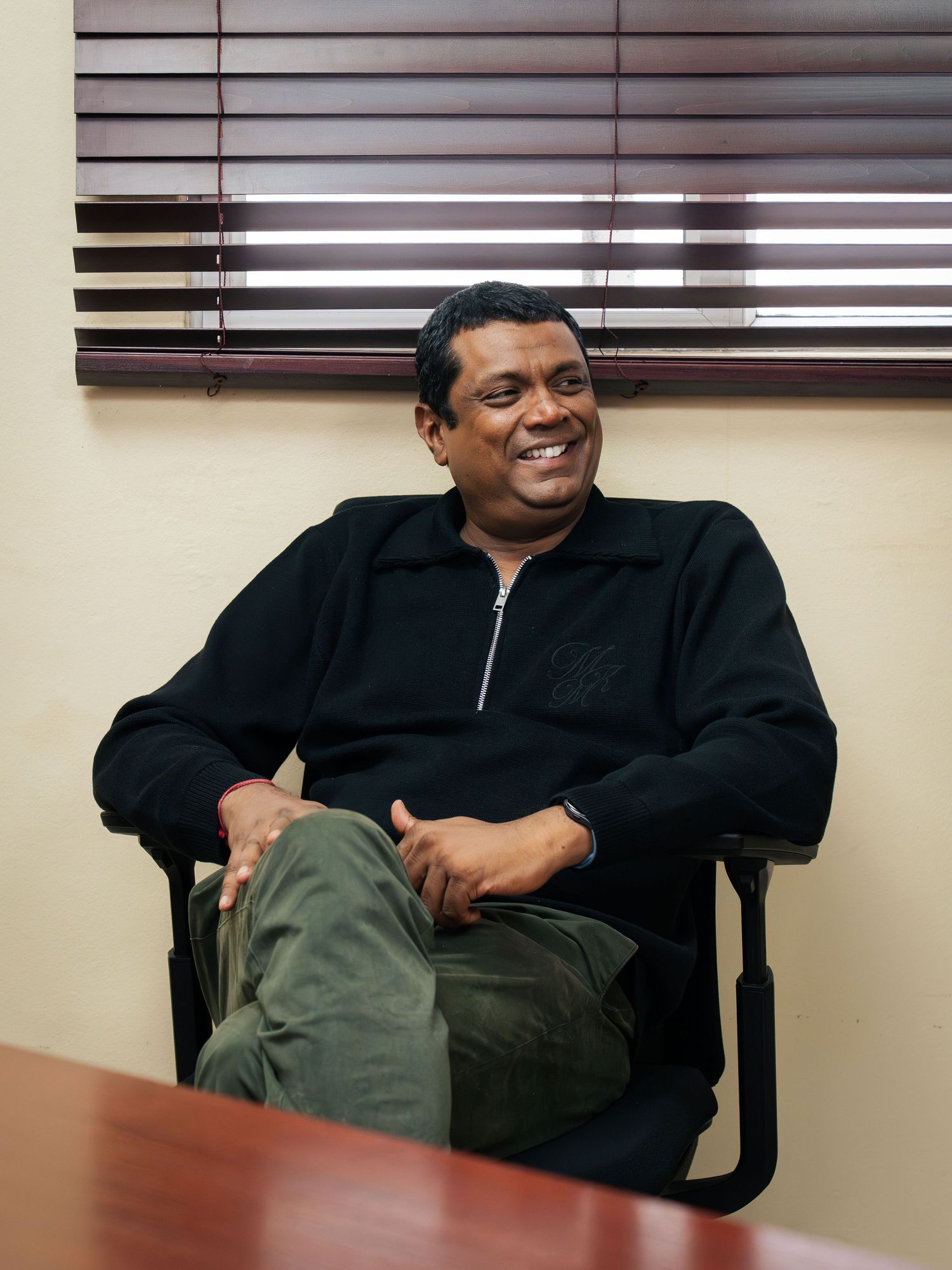 Man in black zip-up sweater and green pants smiling while seated in office chair near wooden table.