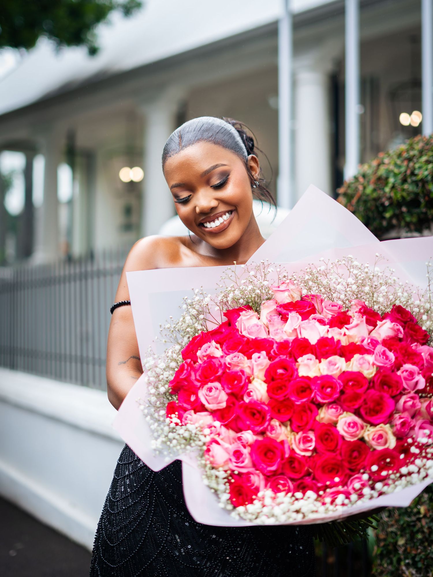 Smiling woman holding a large bouquet of pink and red roses with baby's breath, wearing a black dress outdoors.