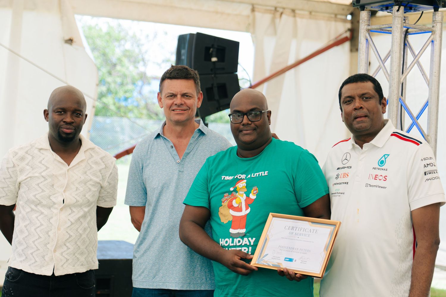 Four men pose together outdoors at an event, one holding a certificate, dressed in casual and branded attire.