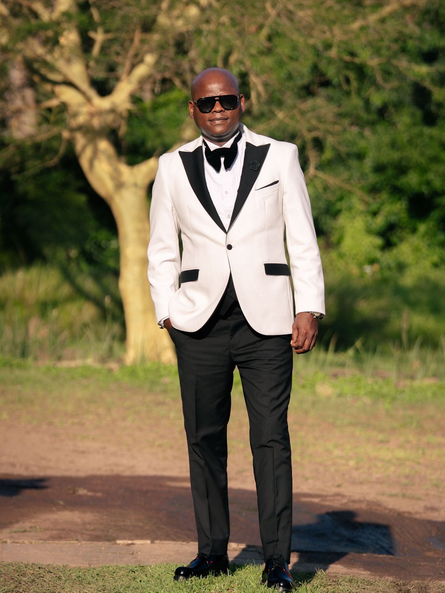 Man in white tuxedo jacket with black lapels, bow tie, and sunglasses standing outdoors near trees.