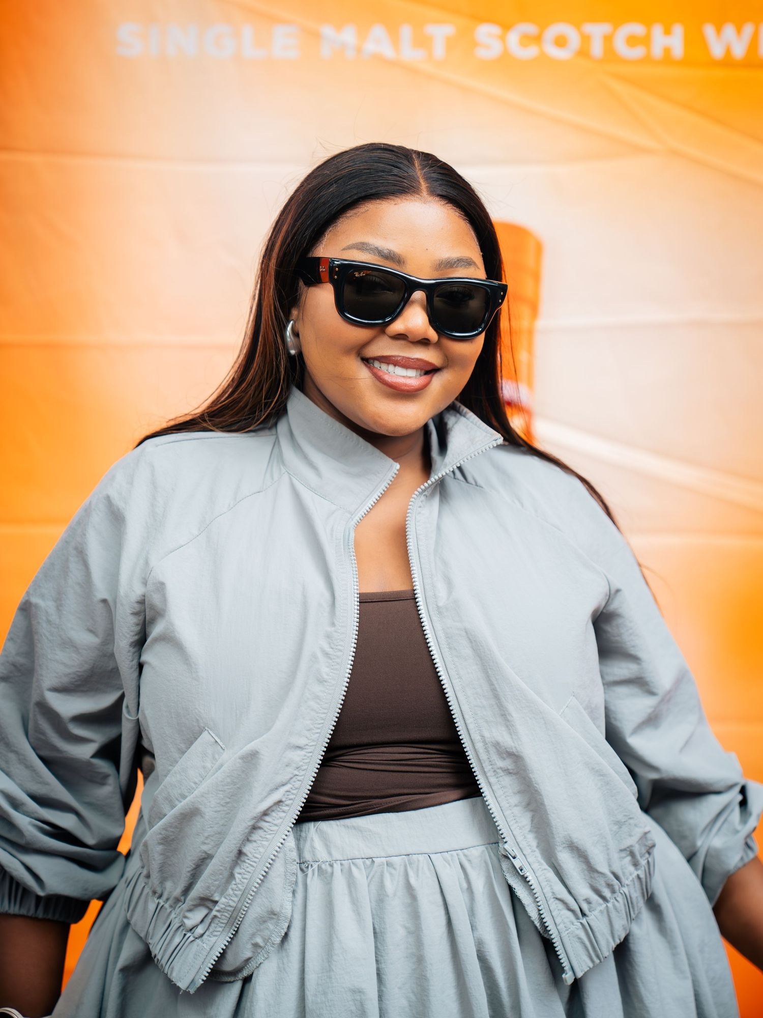 Woman in gray bomber jacket and sunglasses smiling at a Single Malt Scotch whisky event.