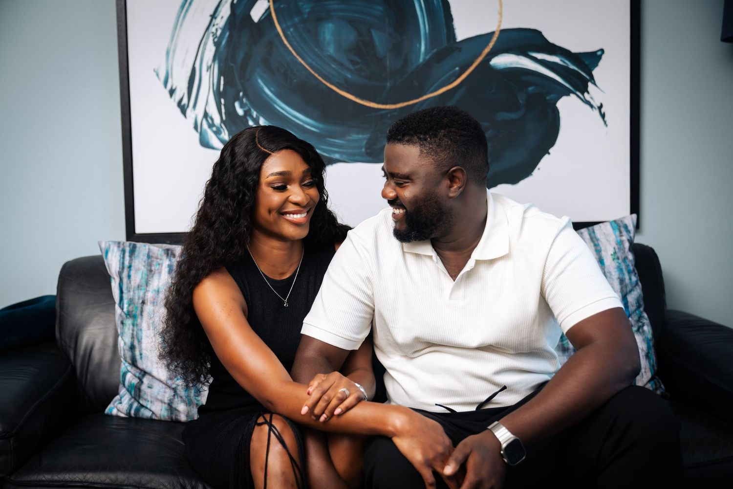 A smiling couple sitting together on a black leather sofa, holding hands in a modern living room.