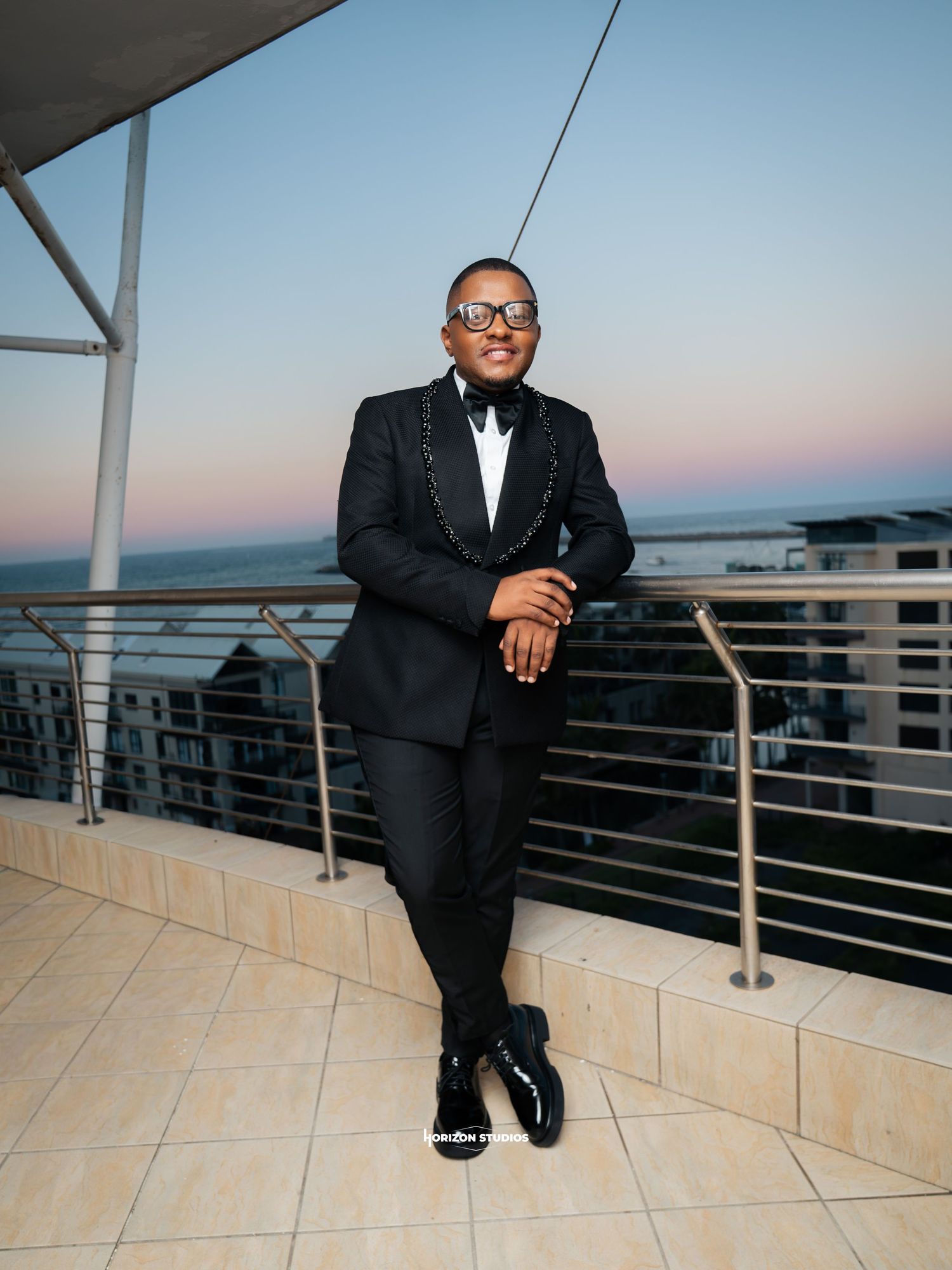 Man in black tuxedo with bow tie poses on rooftop terrace at sunset with ocean view backdrop.