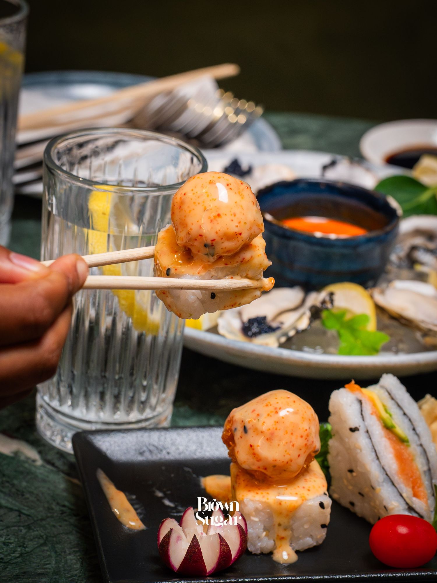 Chopsticks holding a shrimp ball over sushi rice on a black tray, with sauces and oysters in the background.