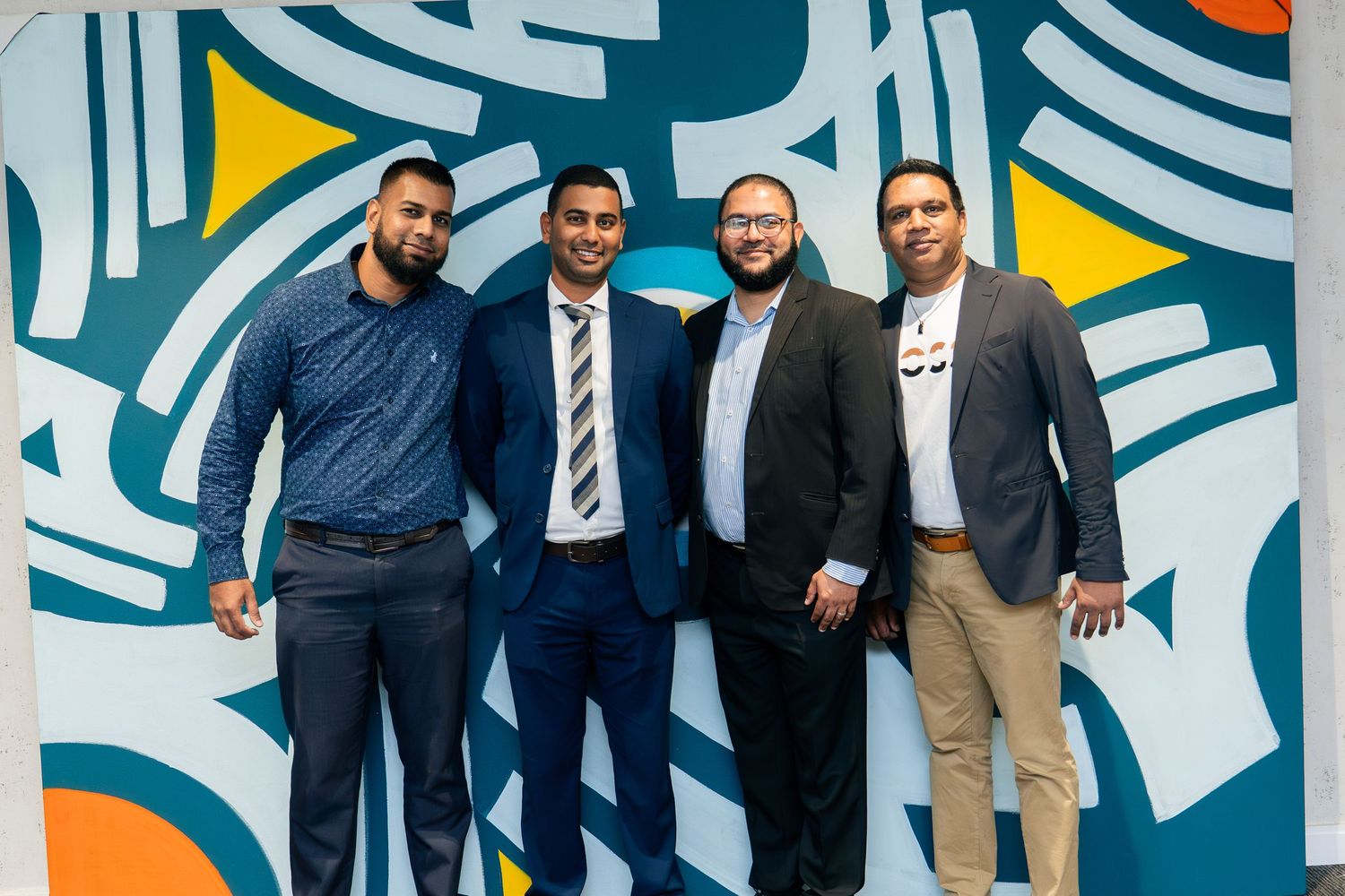 Four men in formal attire smiling together in front of a colorful abstract mural with blue, orange, and white tones.