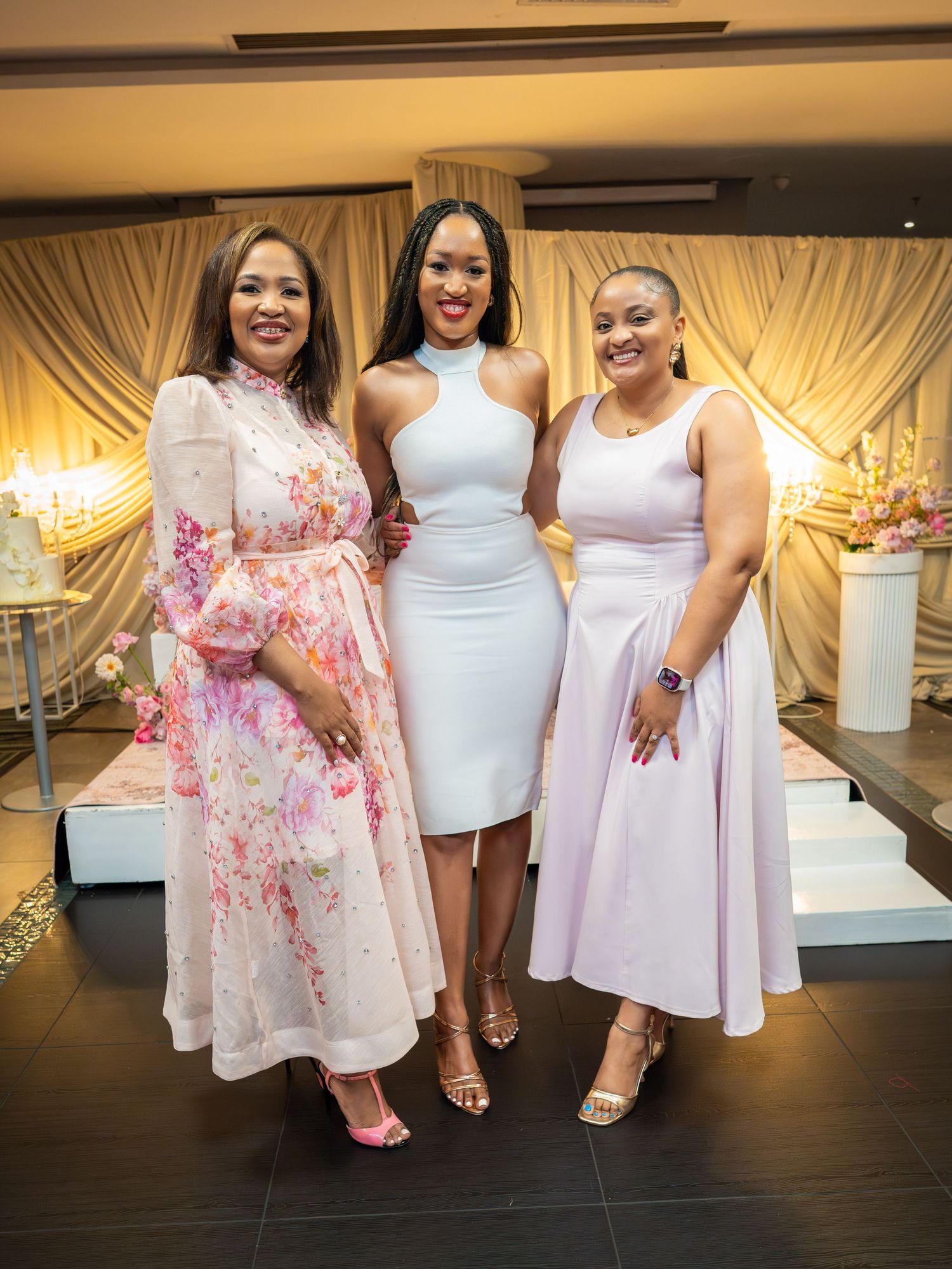 Three elegant women posing together at a formal event, wearing white and pastel dresses in a decorated venue.