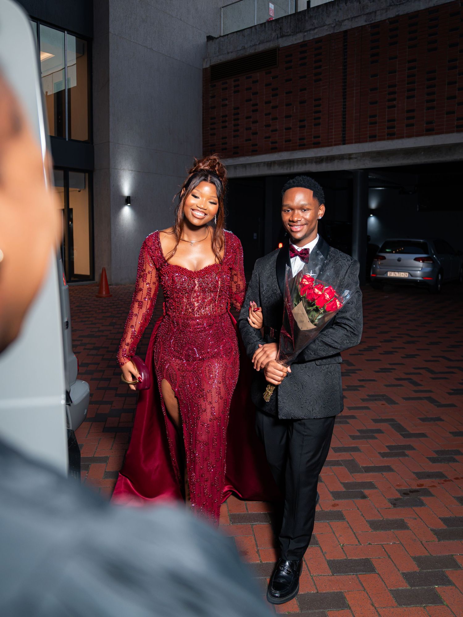 Couple dressed in elegant formal attire, woman in red lace gown and man in suit holding roses, posing at a prom event.