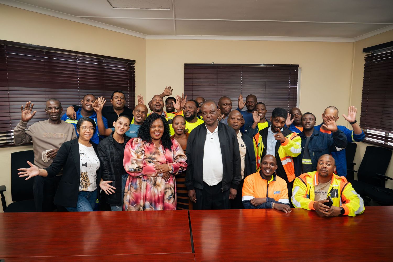 Large group of diverse professionals posing together in a conference room, smiling and waving at the camera.