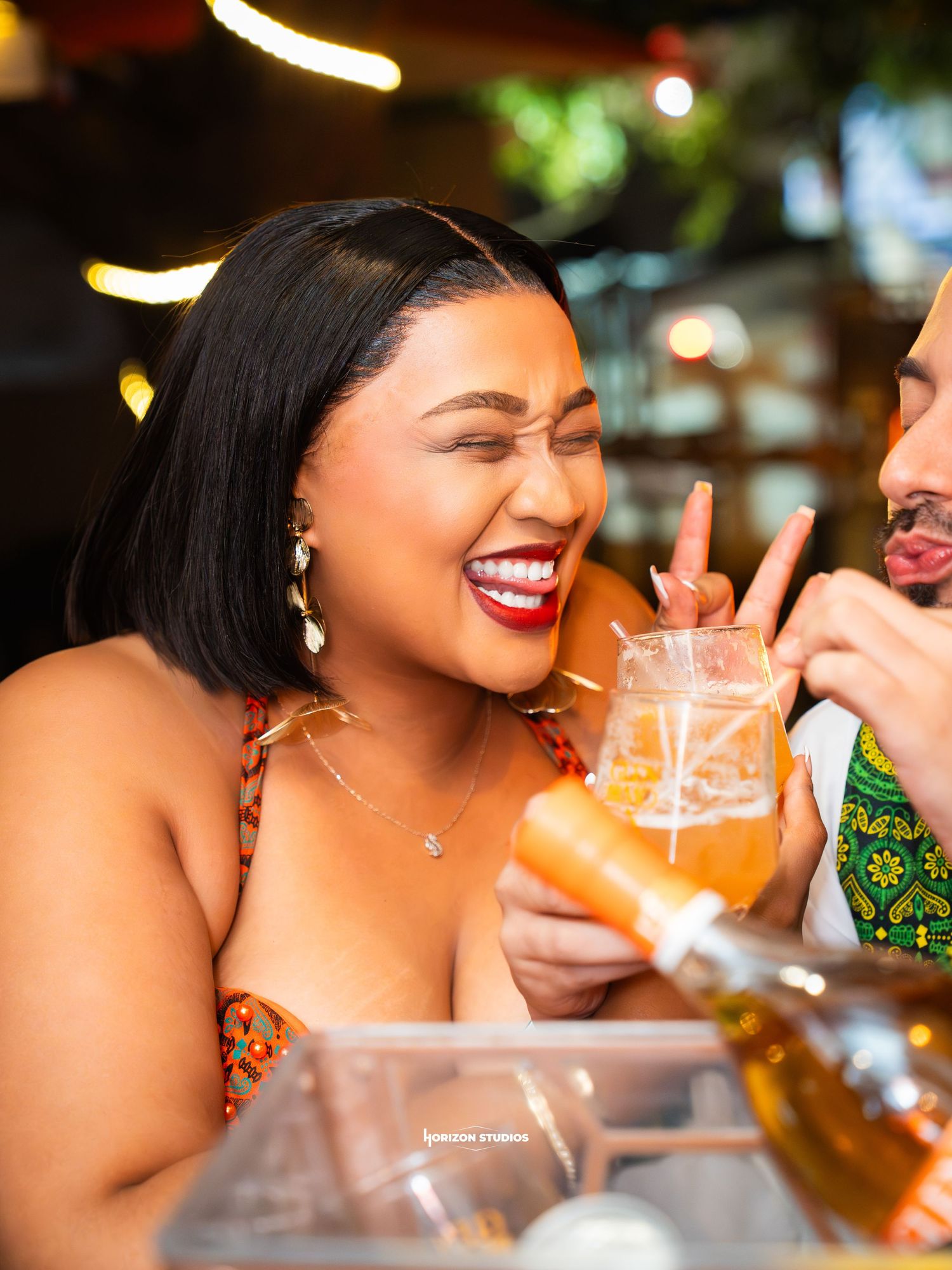 A woman laughs joyfully while holding a cocktail at a bar, wearing a colorful dress and red lipstick.