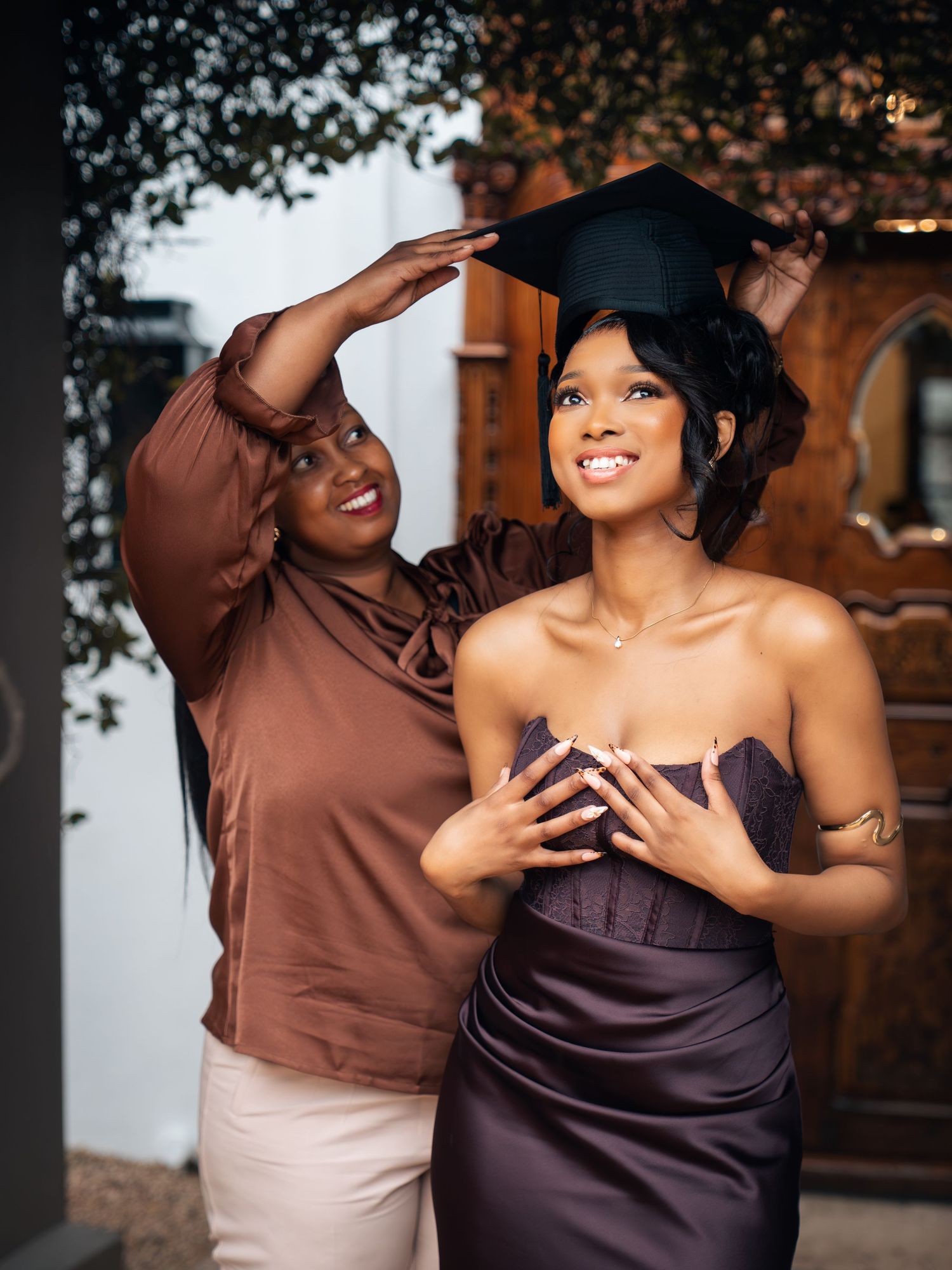 A proud mother places a graduation cap on her smiling daughter wearing an elegant strapless purple gown.
