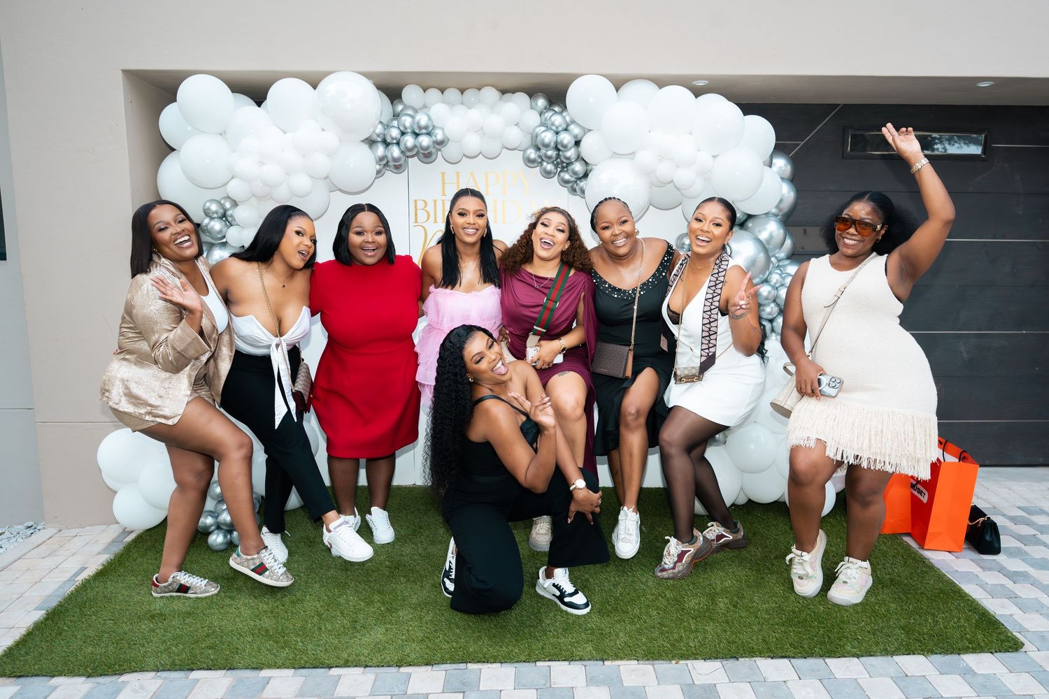 Group of women posing and celebrating at a party with white balloon arch backdrop on green turf.