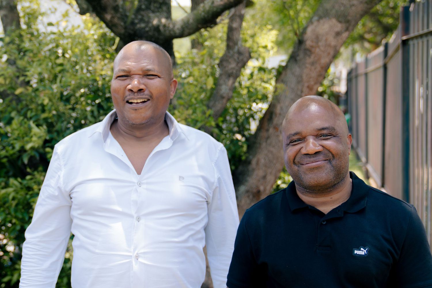 Two smiling men standing outdoors near trees, one in a white shirt and one in a dark Puma polo shirt.