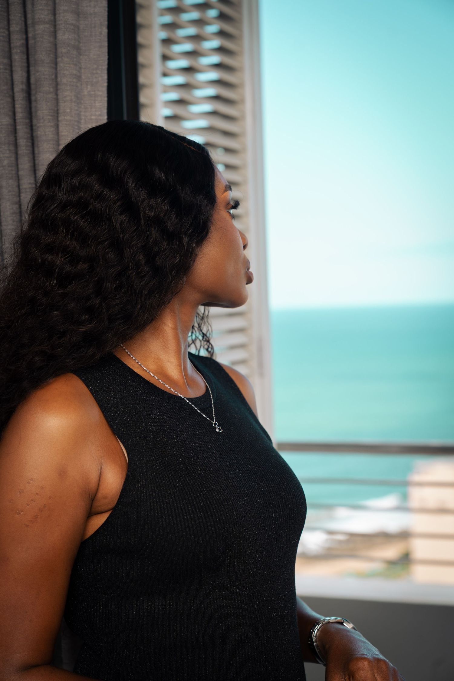 A Black woman in a black sleeveless dress gazes out a window with an ocean view, wearing a silver necklace.