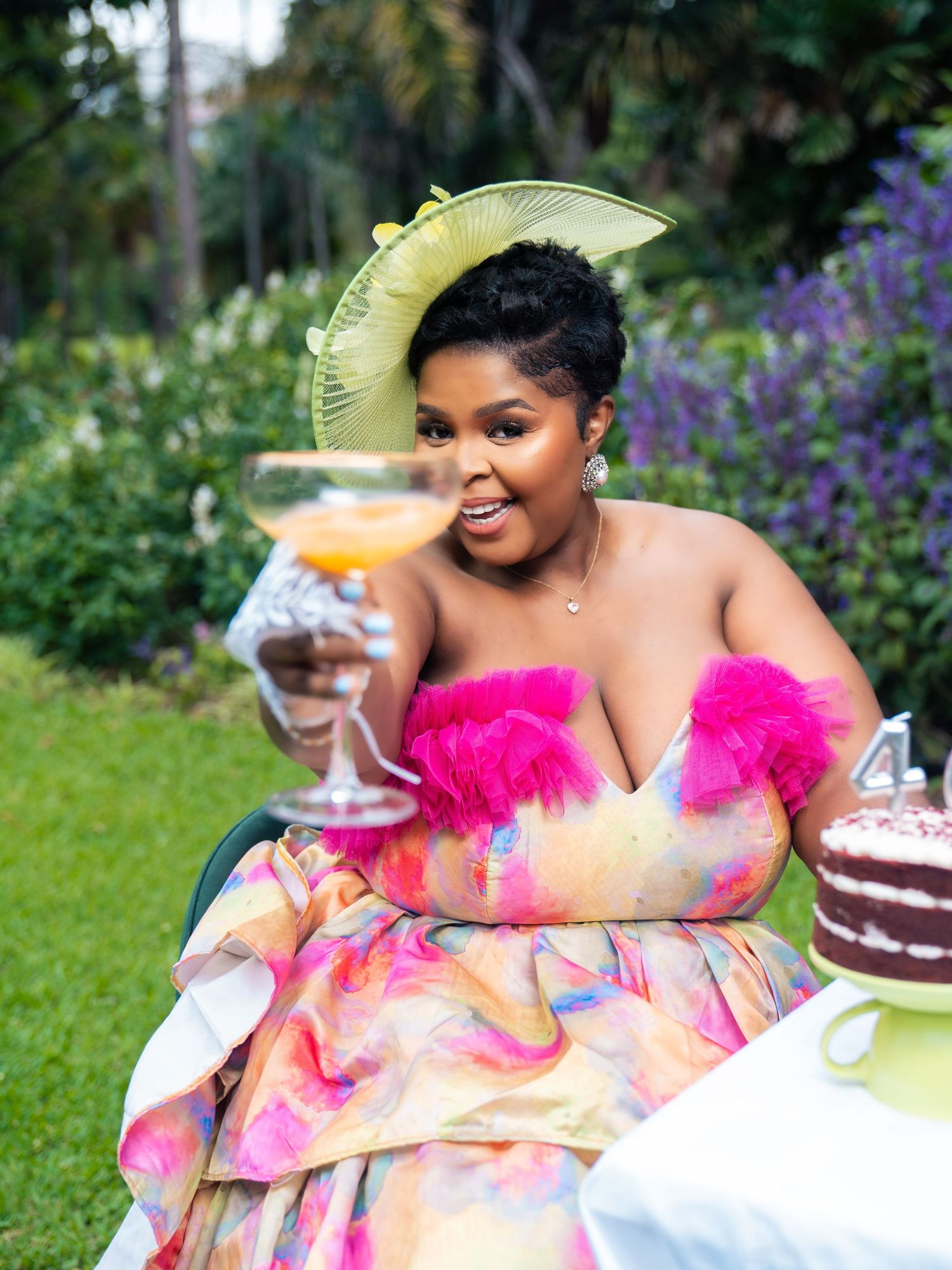 Woman in colorful gown and green hat holds oversized cocktail glass outdoors near birthday cake.