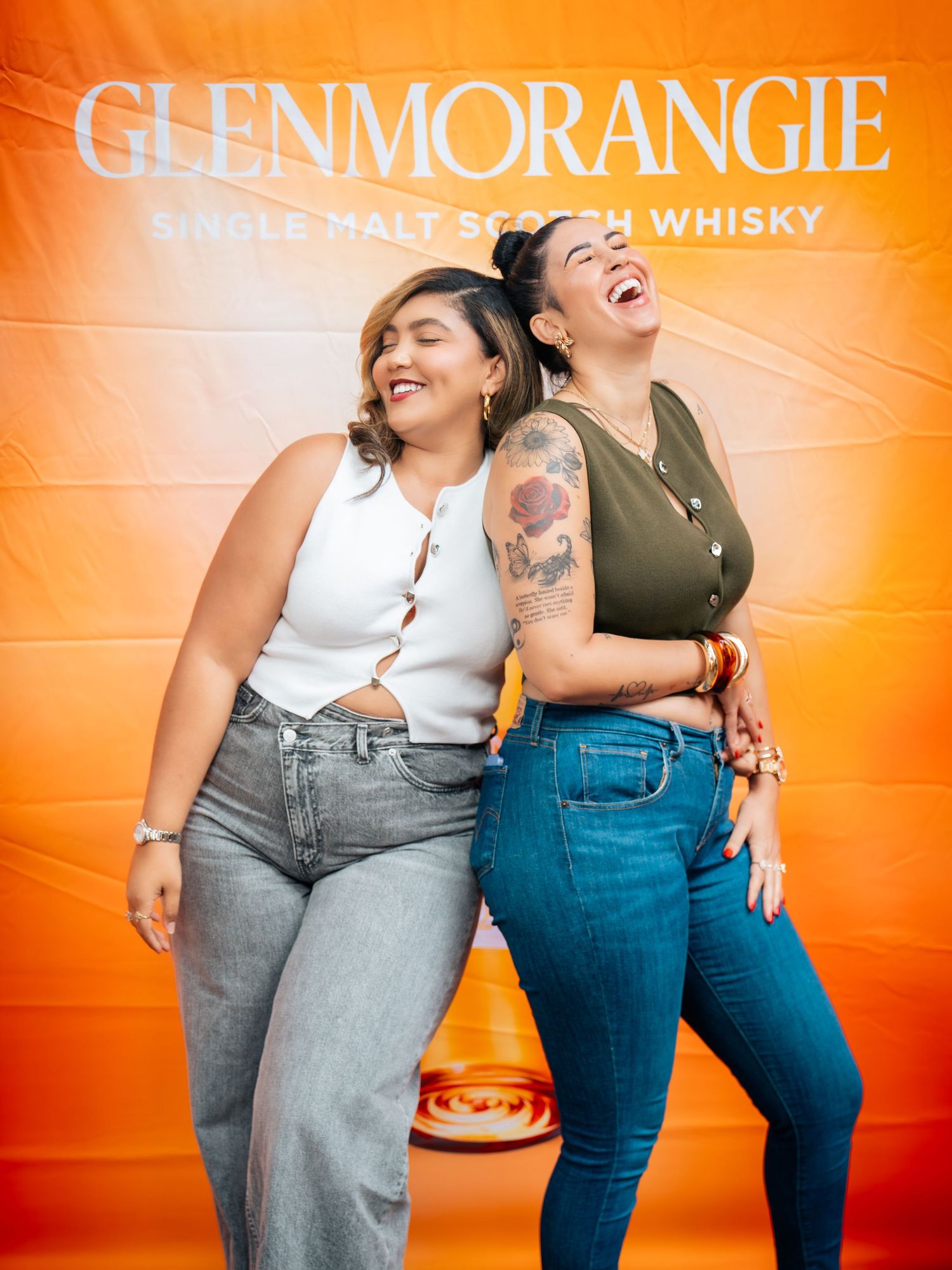 Two women laughing and posing in front of a Glenmorangie Single Malt Scotch Whisky orange backdrop.