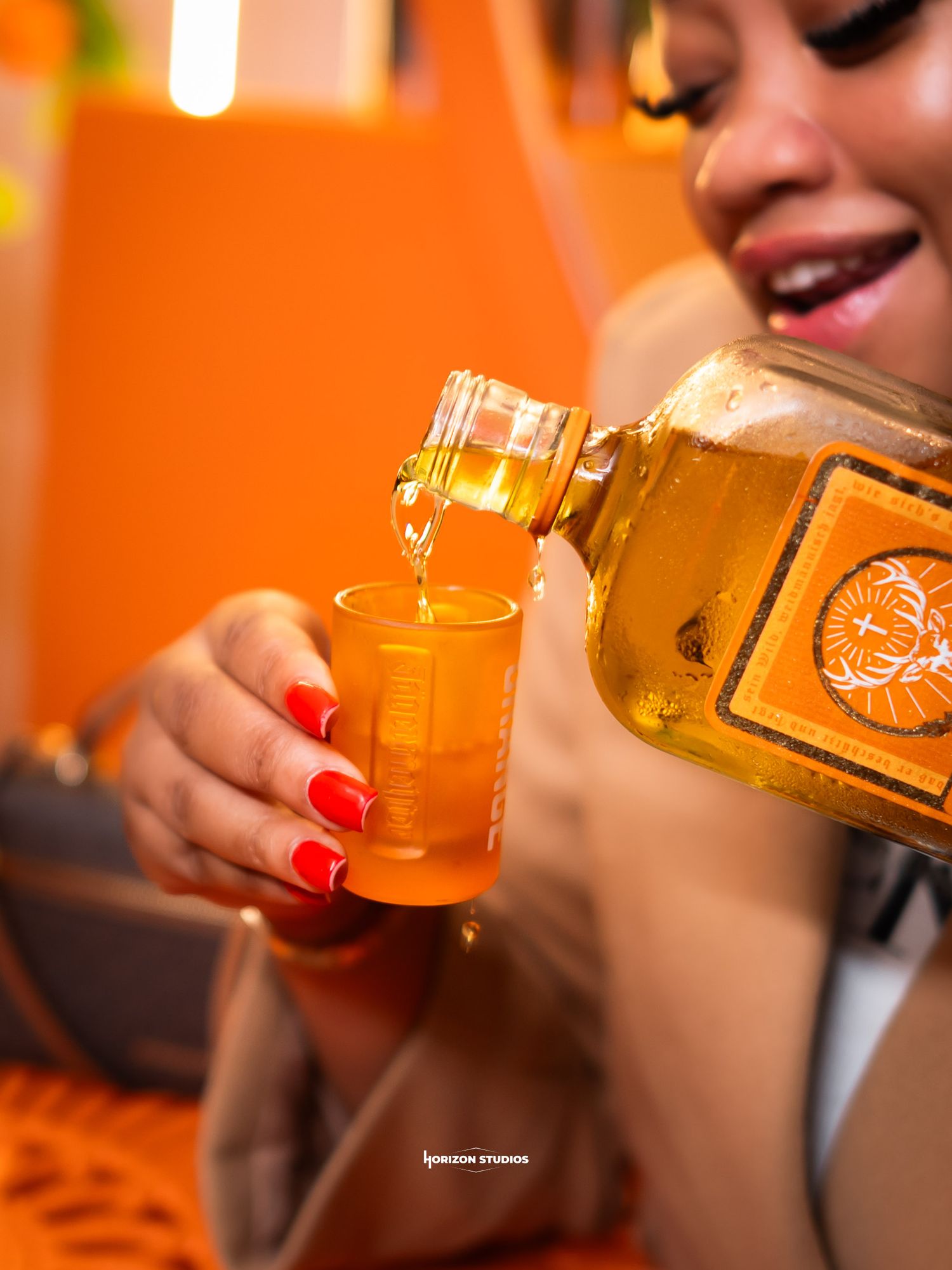 Woman with red nails pouring amber tequila from a decorative bottle into a shot glass against an orange background.