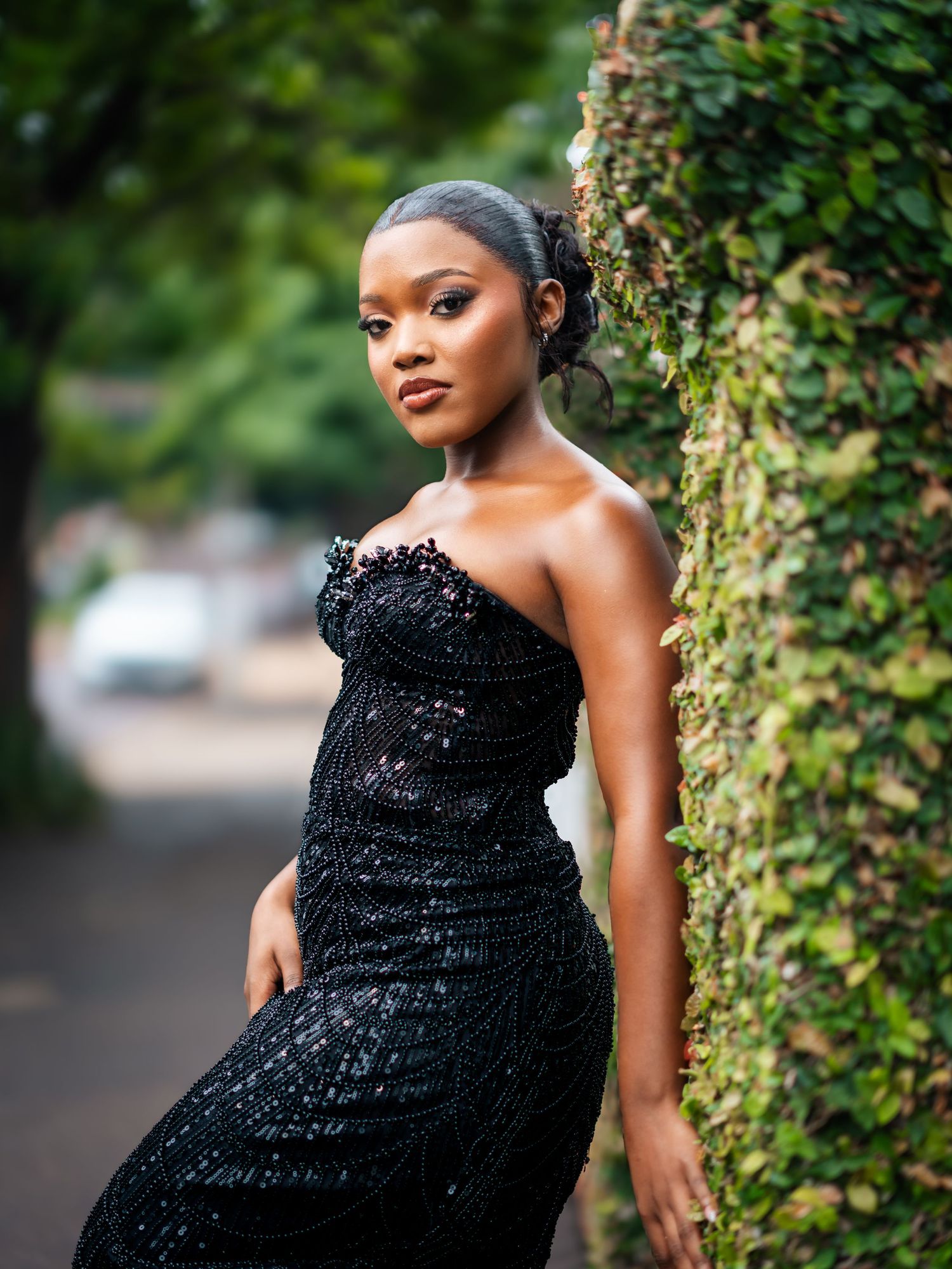 Young Black woman in elegant black sequin strapless gown posing beside ivy-covered wall outdoors.