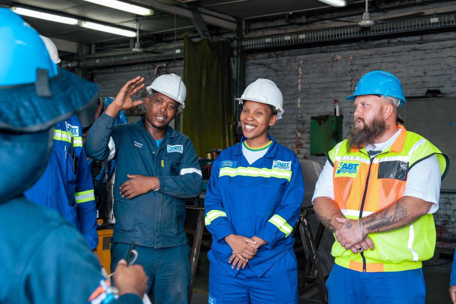 Industrial workers in hard hats and safety gear smile during a meeting inside a factory facility.