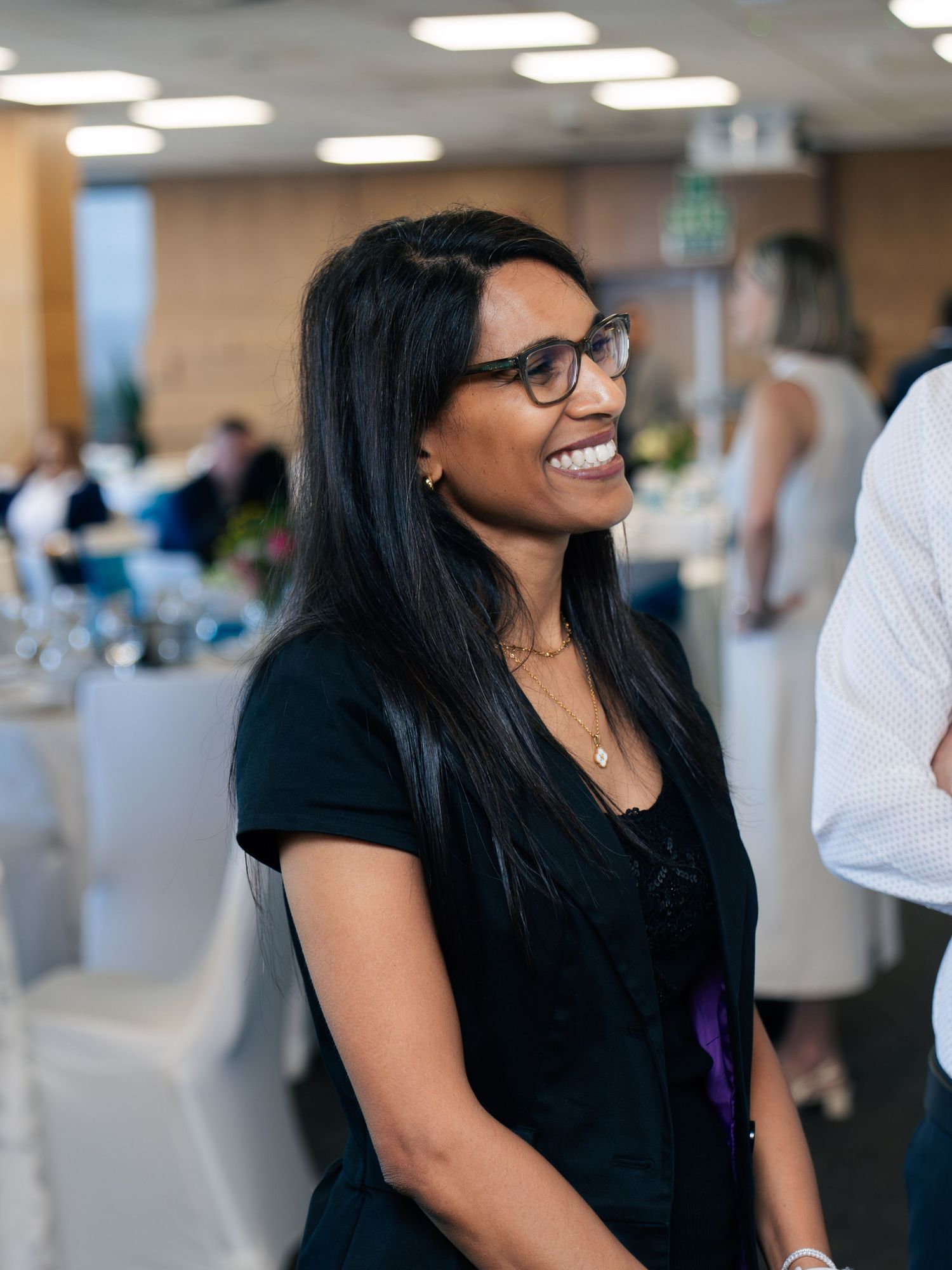 Smiling woman with glasses and long black hair at an indoor event, wearing a black outfit.