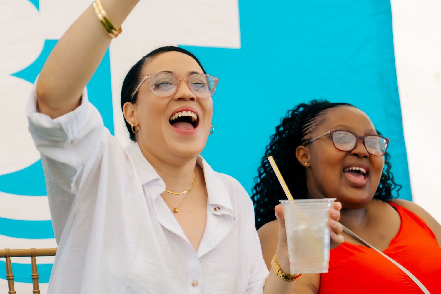 Two joyful women laughing and celebrating outdoors, one in white raising her arm, the other in orange holding a drink.