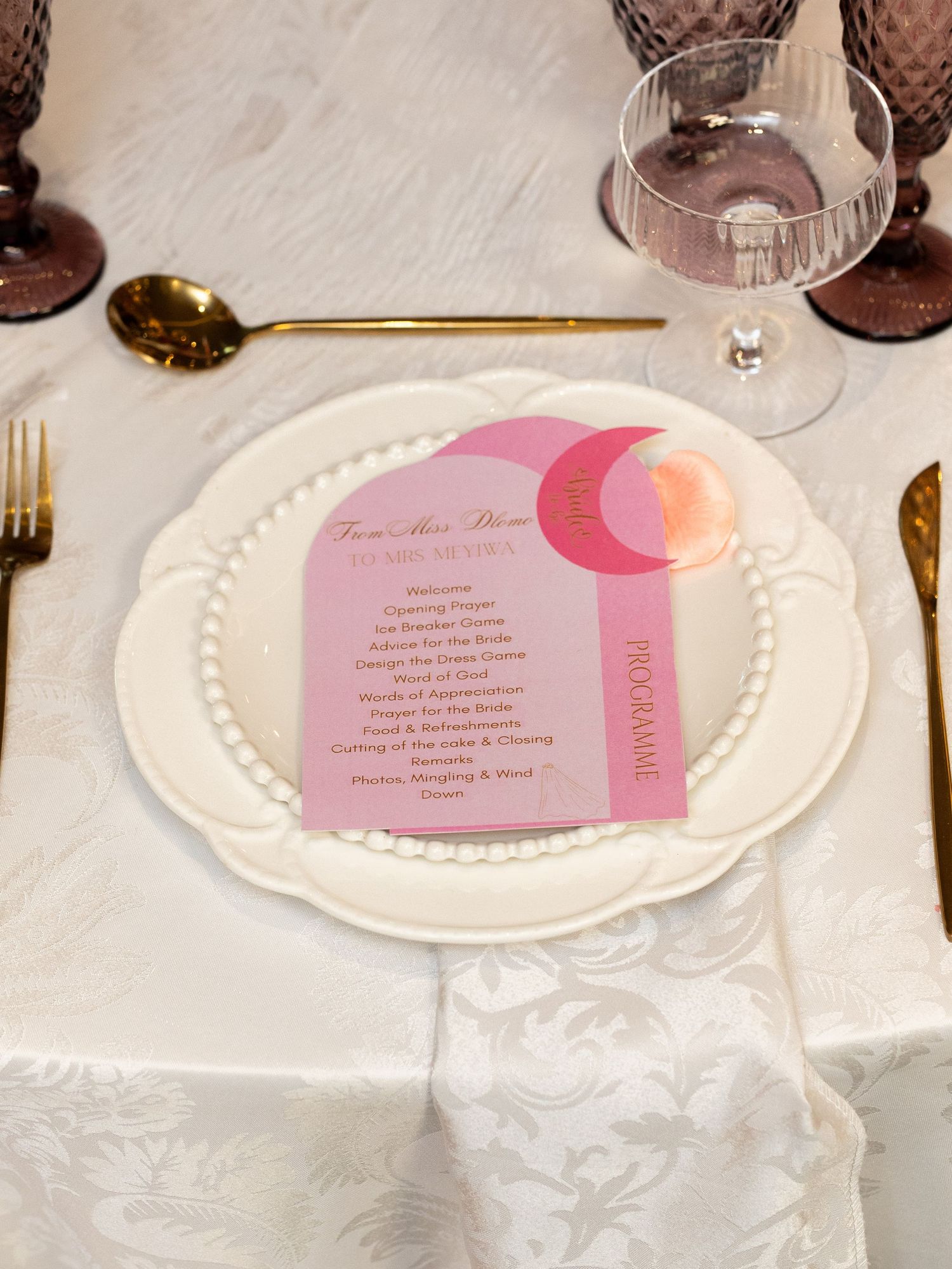 Elegant bridal shower place setting with pink program card, gold cutlery, and purple glassware.