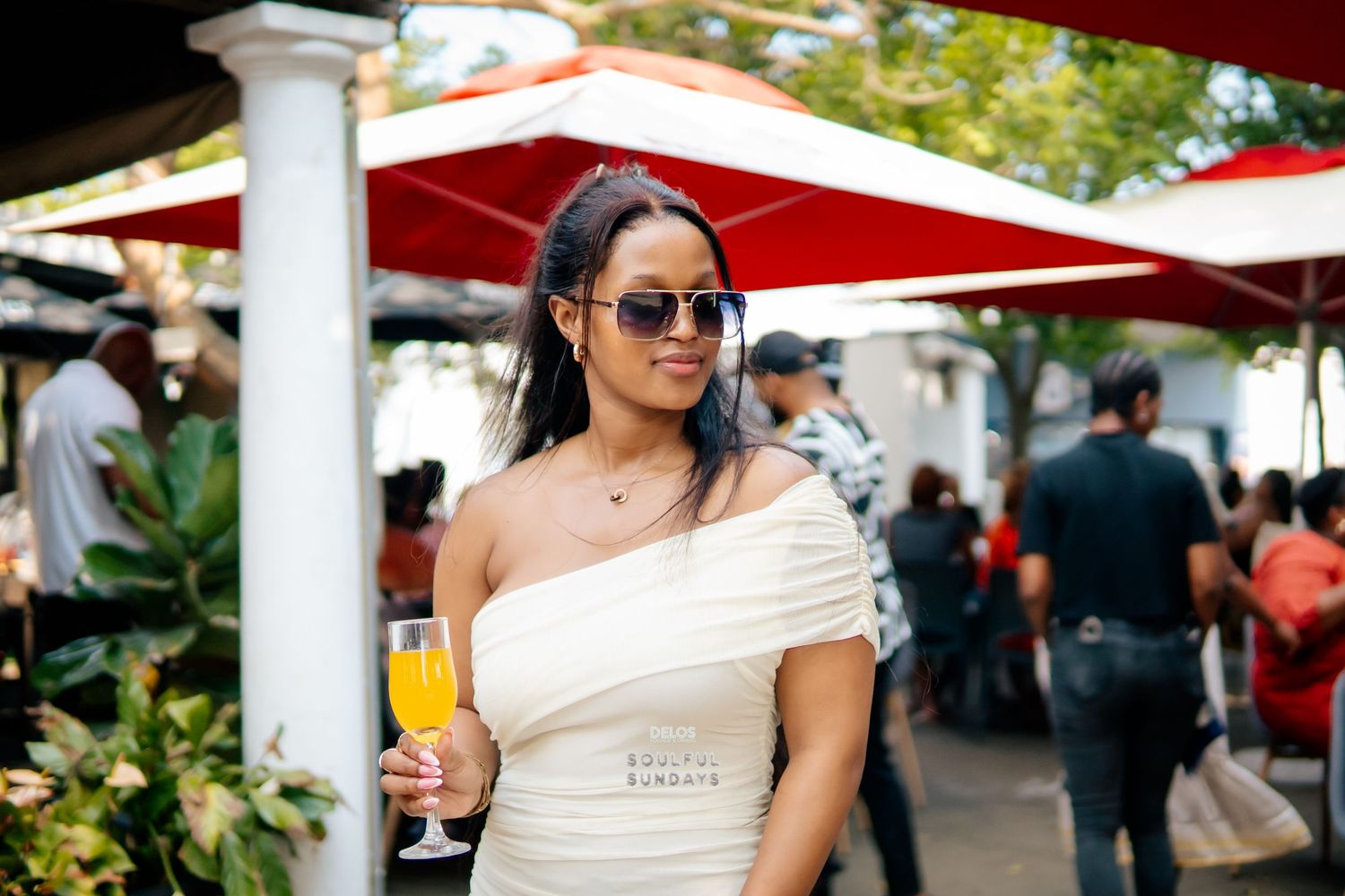 Woman in white off-shoulder dress holding mimosa at outdoor brunch event, wearing sunglasses and gold necklace.