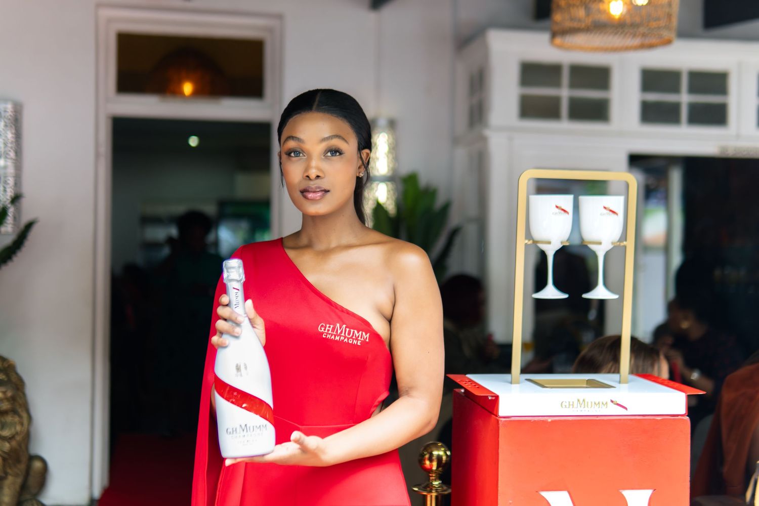 Woman in red one-shoulder dress holding a white bottle at an elegant event with wine glasses displayed.