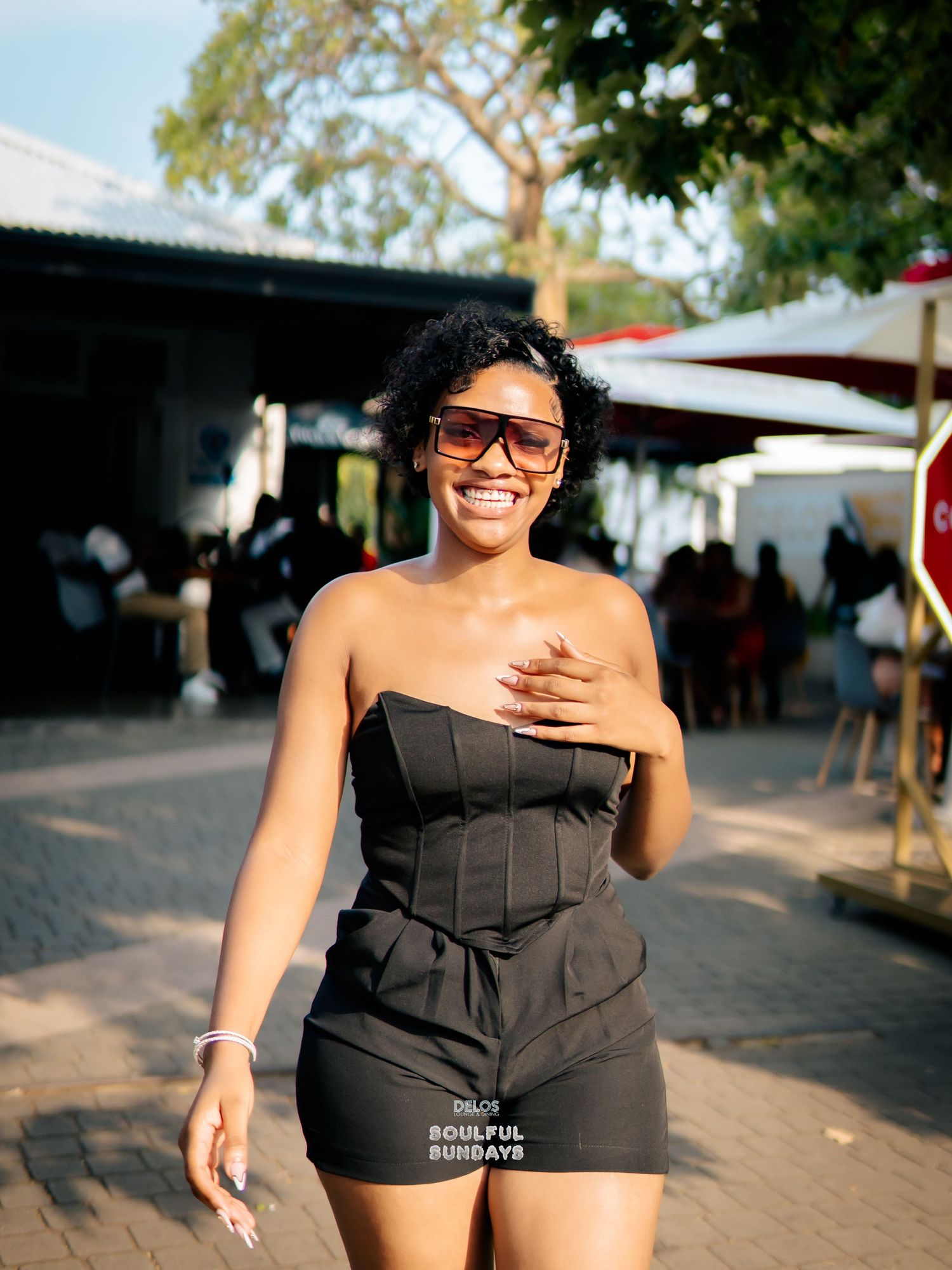 A smiling woman in a black strapless corset outfit poses outdoors at a social event on a sunny day.