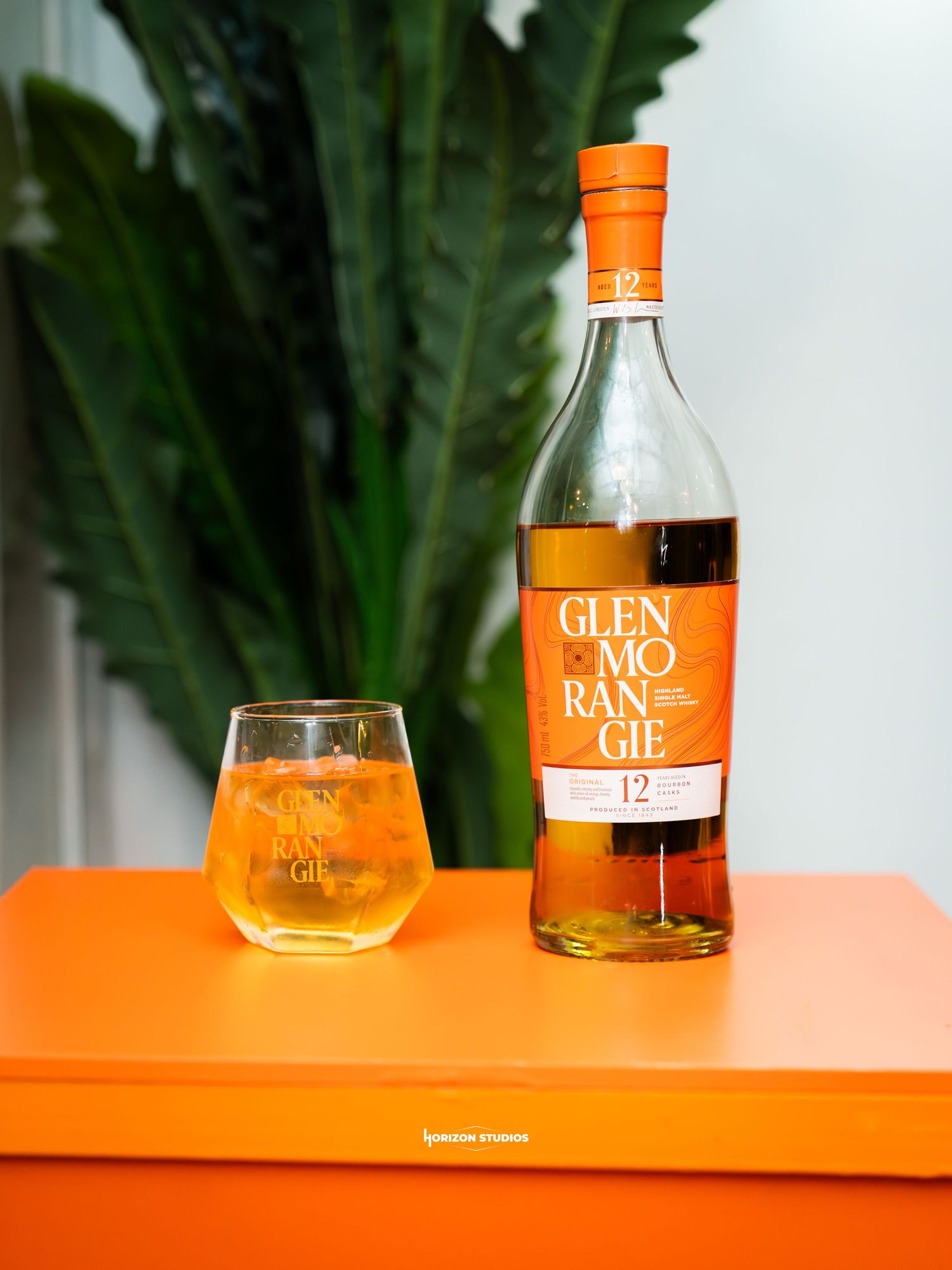 Glenmorangie 12 year old Scotch whisky bottle beside a filled glass on an orange surface with tropical plants.