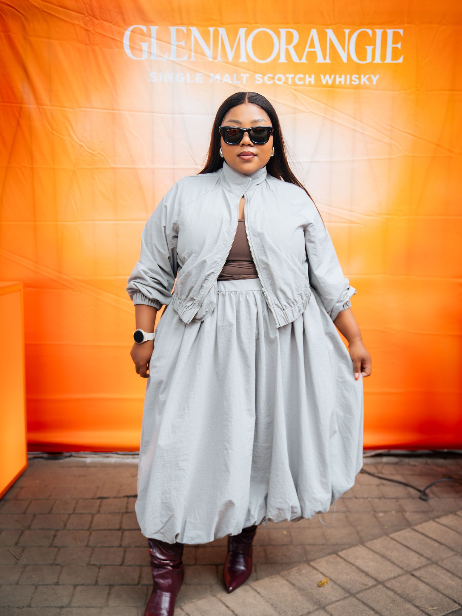 Woman in gray midi skirt and jacket with dark sunglasses poses before Glenmorangie Scotch Whisky orange backdrop.