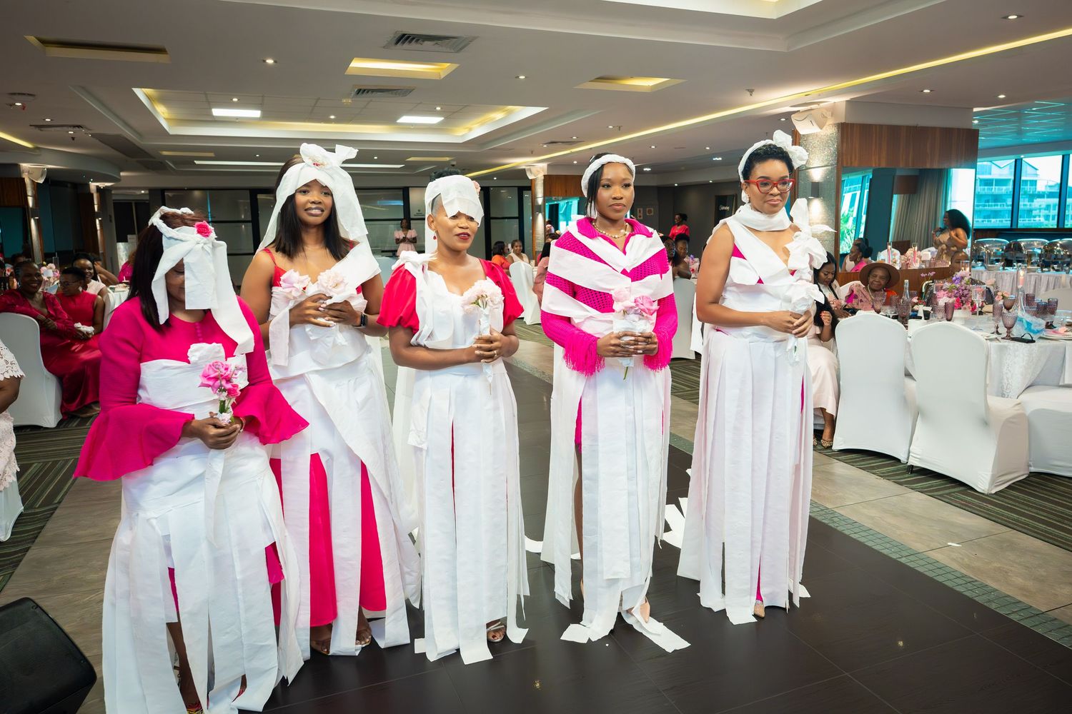 Women in white and pink outfits with head wraps standing in a decorated banquet hall during a cultural ceremony.