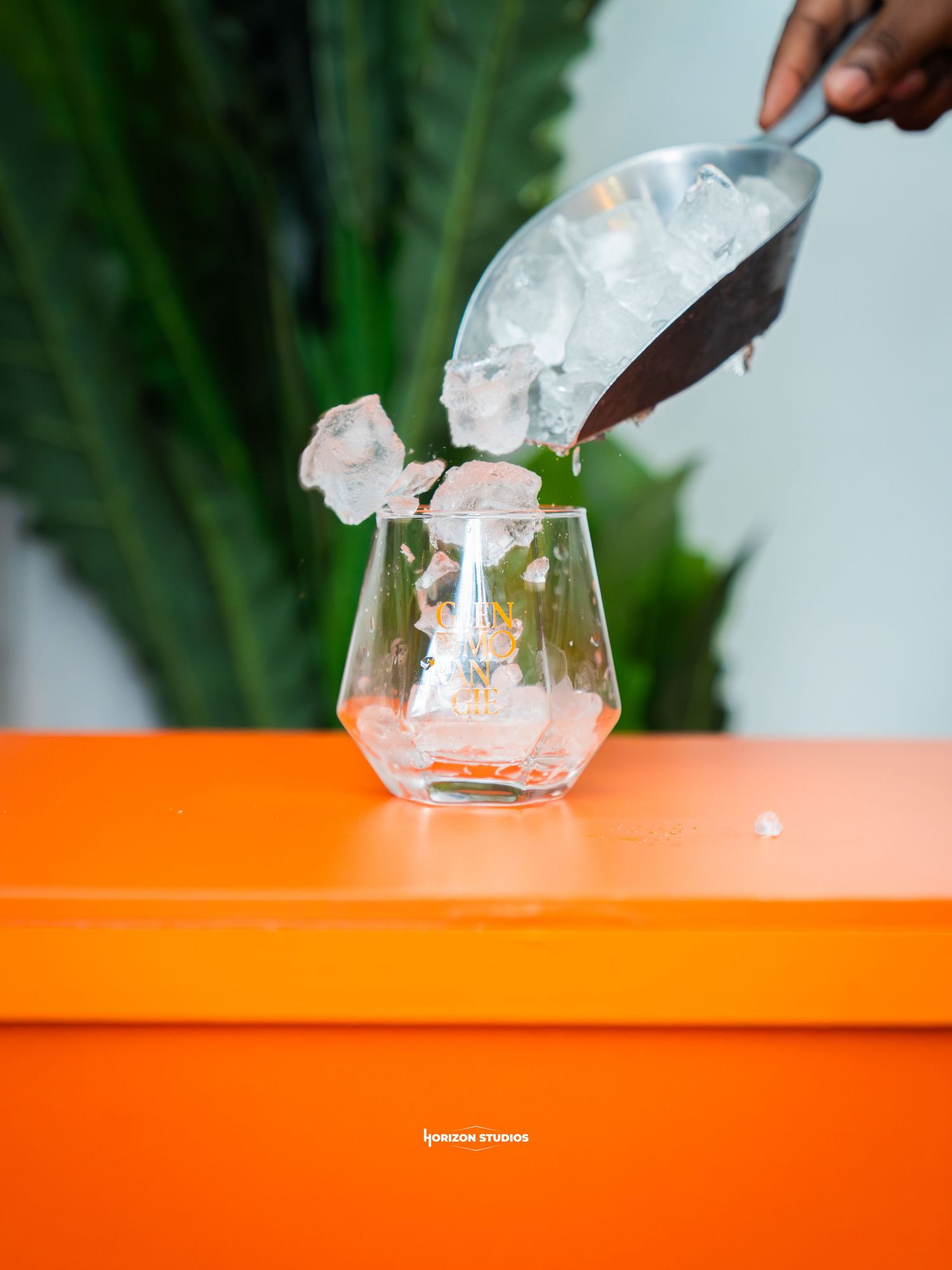 Hand pouring ice cubes from a metal scoop into a stemless glass on a bright orange table.