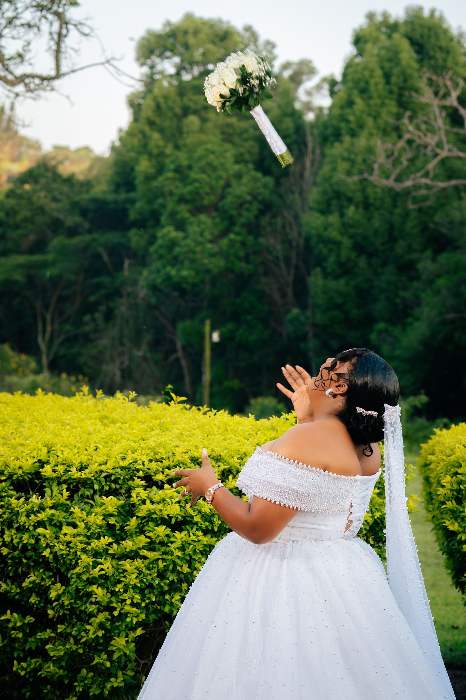 Bride in white off-shoulder gown tossing bouquet in lush green garden setting.